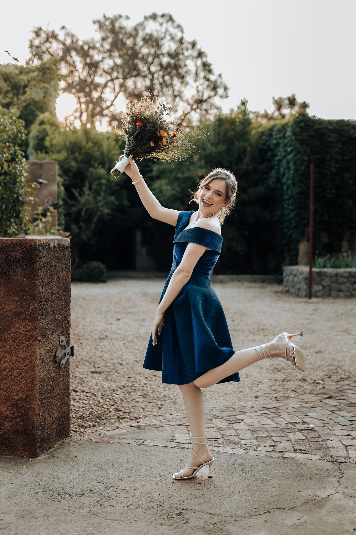 Bridesmaid in navy dress playfully poses with bouquet raised while standing on one leg.