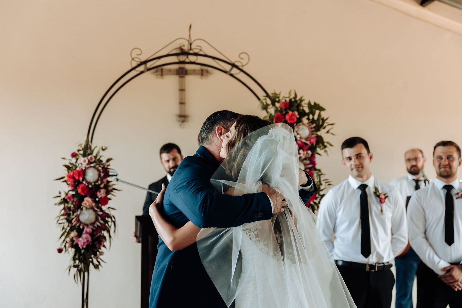 Wedding ceremony under a floral arch with pink and red roses in an indoor venue.