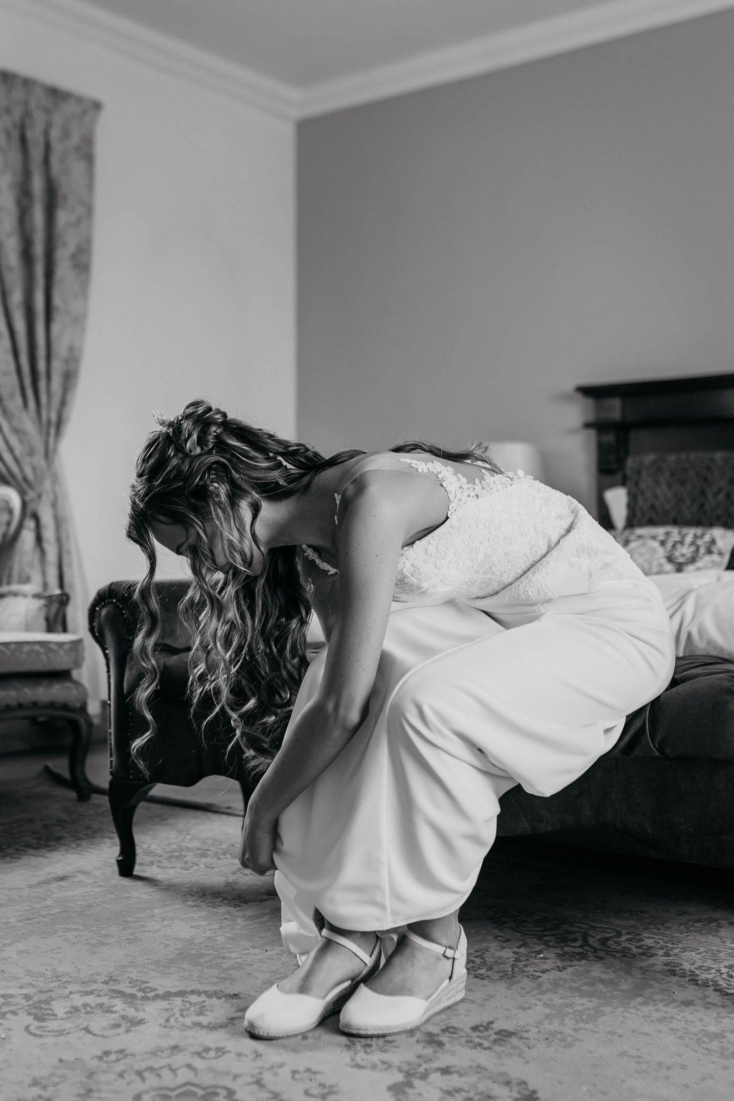 A bride in a white wedding gown adjusts her shoe in an elegant bedroom setting.