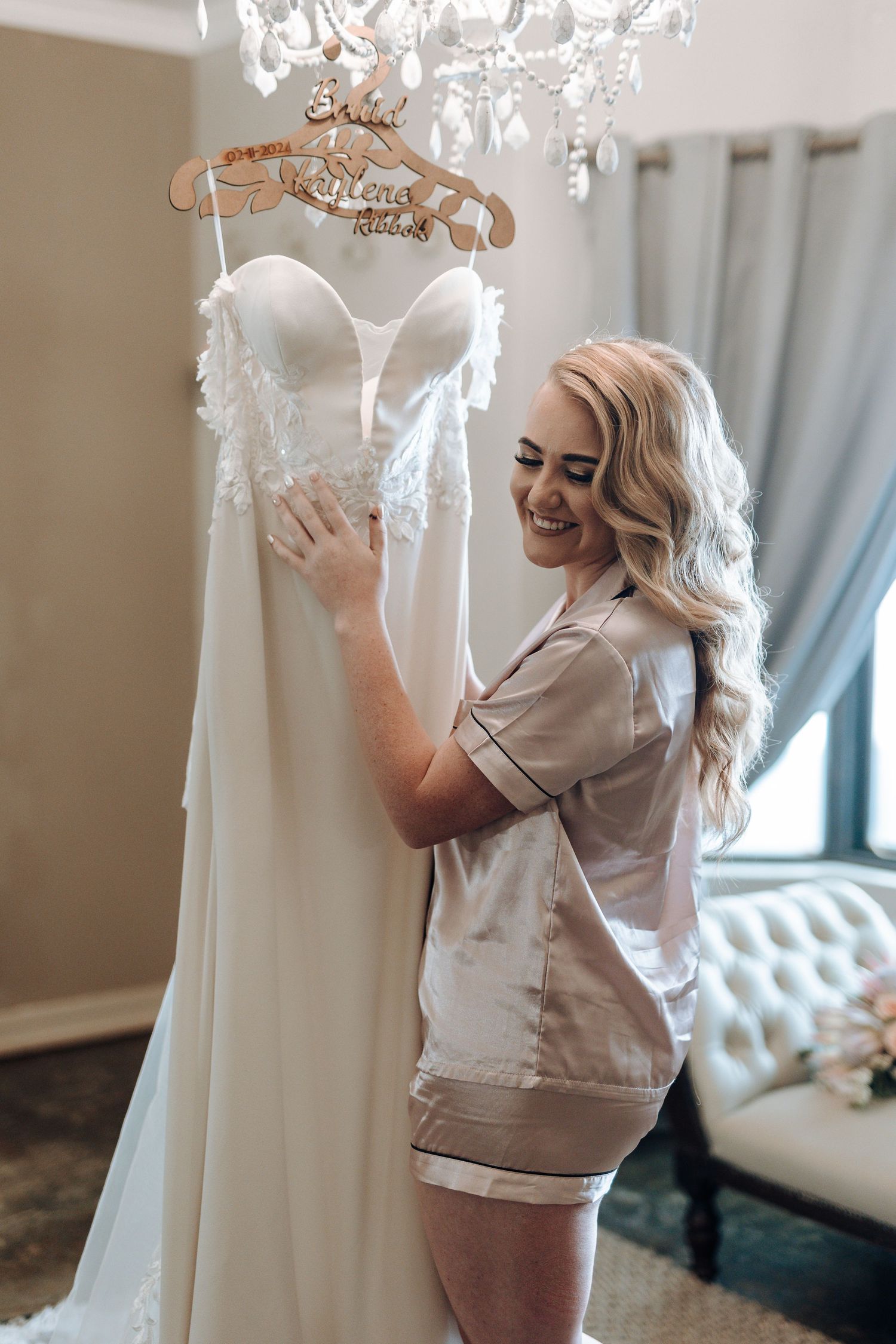 Bride in getting ready attire admires white wedding gown hanging from crystal chandelier.