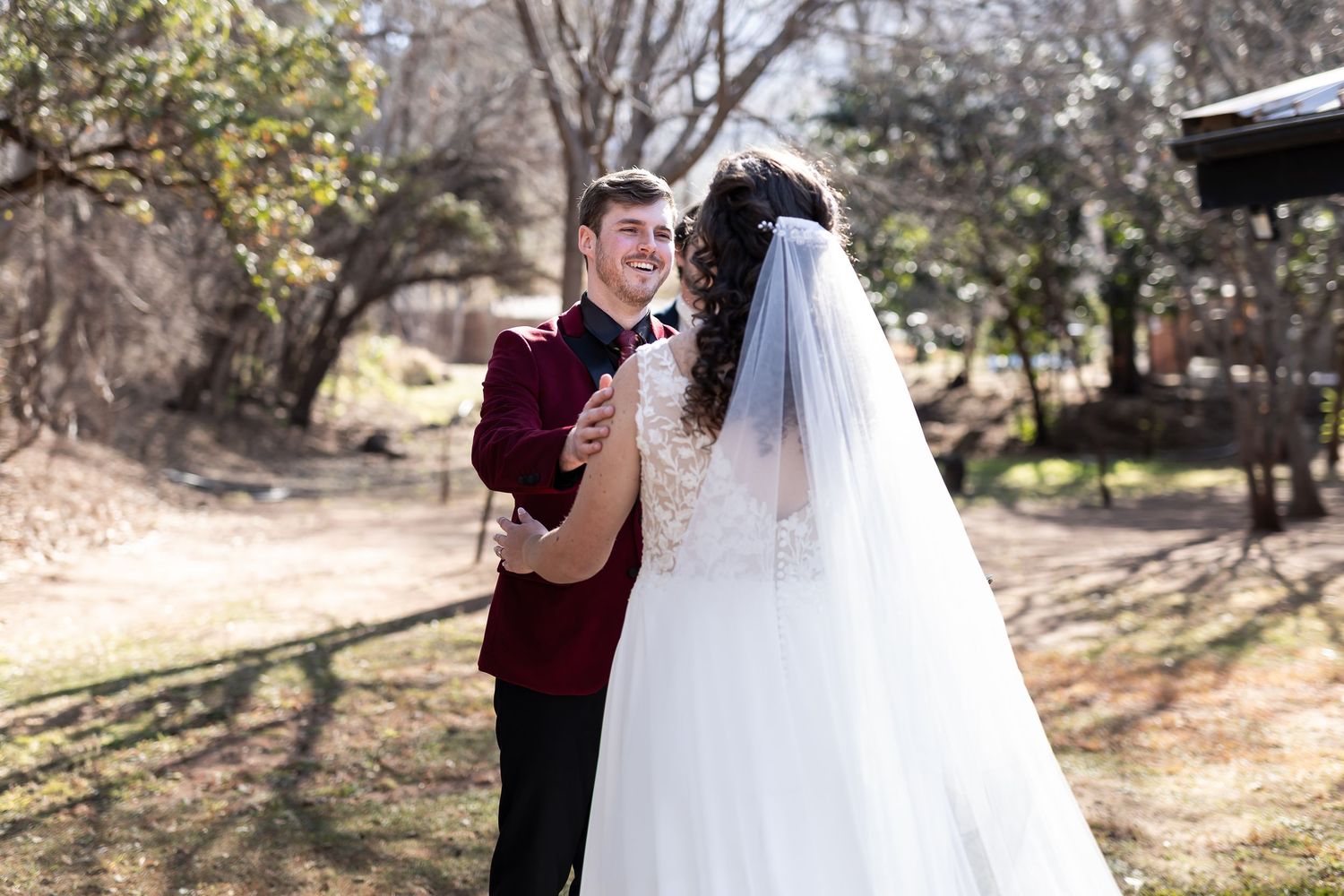 A bride in a white wedding dress with a long veil embraces someone wearing a burgundy suit in a sunlit outdoor setting.