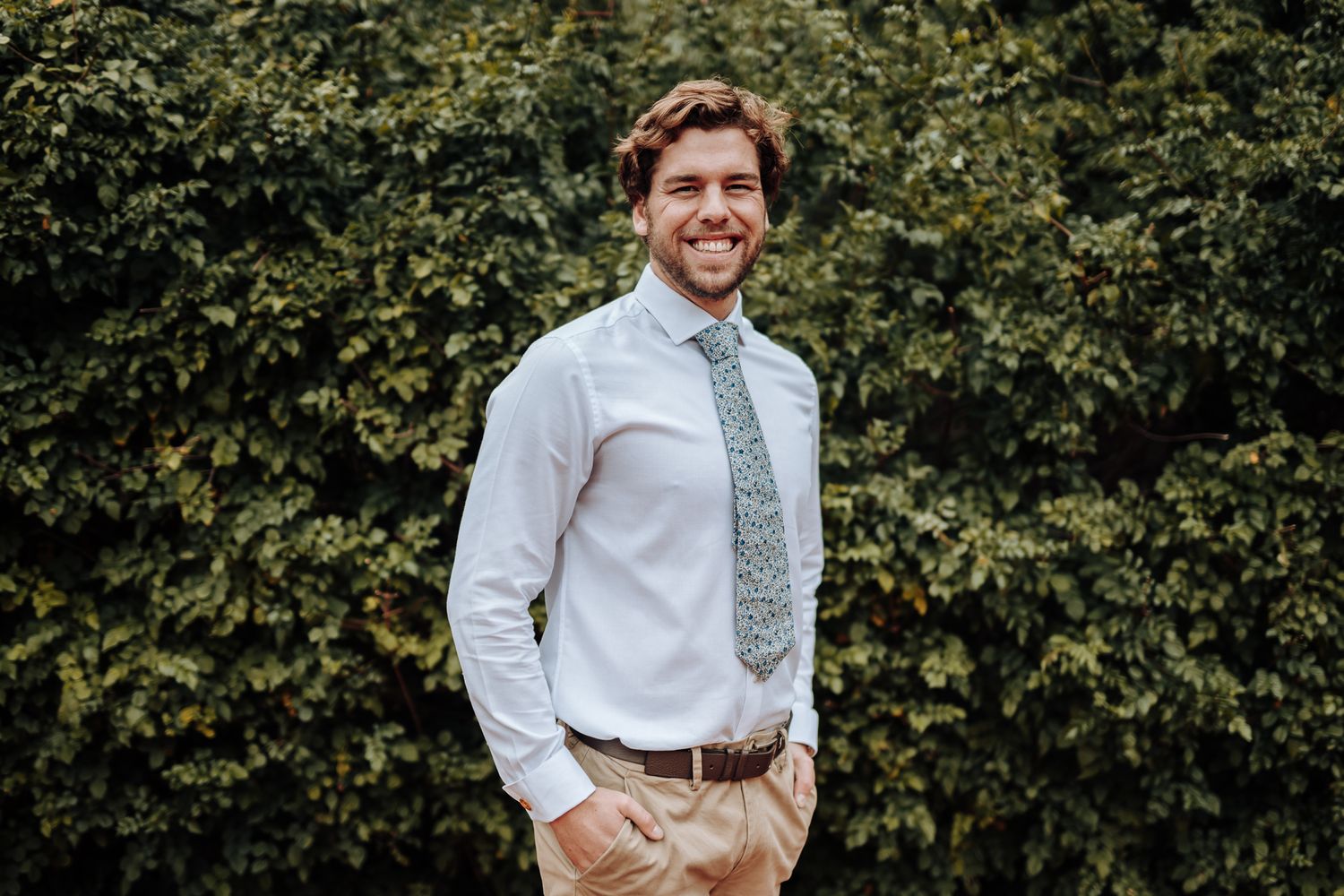 Wedding party member in crisp white shirt and patterned tie stands against green shrubbery.