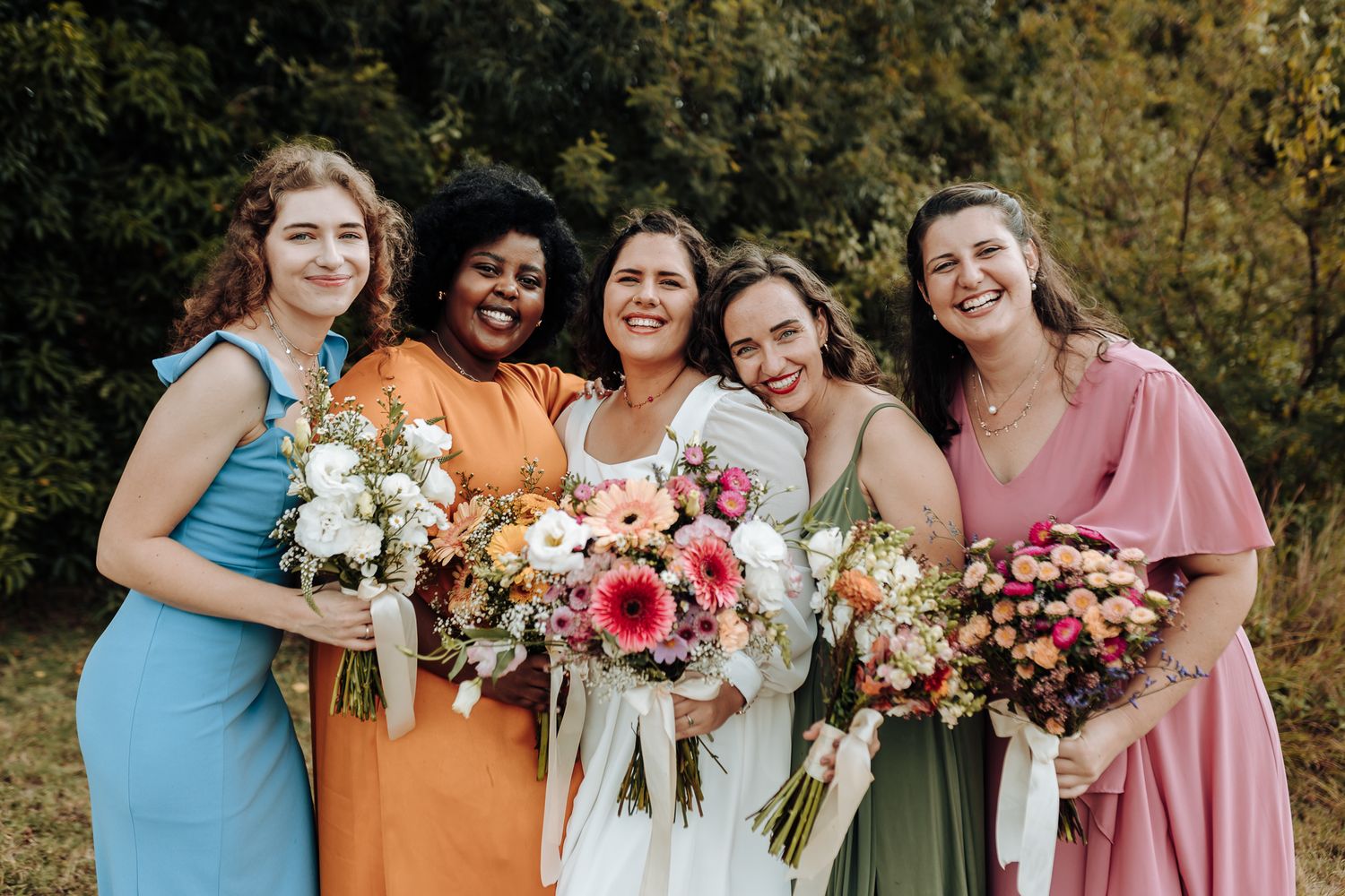 Wedding party in mismatched dress colors holding vibrant floral bouquets in outdoor setting.