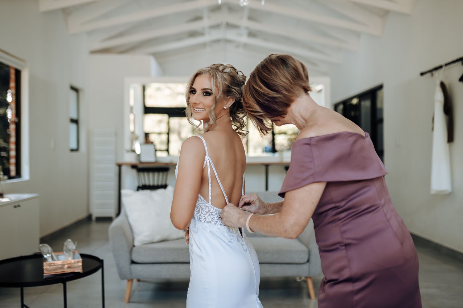 Person in mauve dress helps button up a delicate white lace wedding gown in bright modern room.