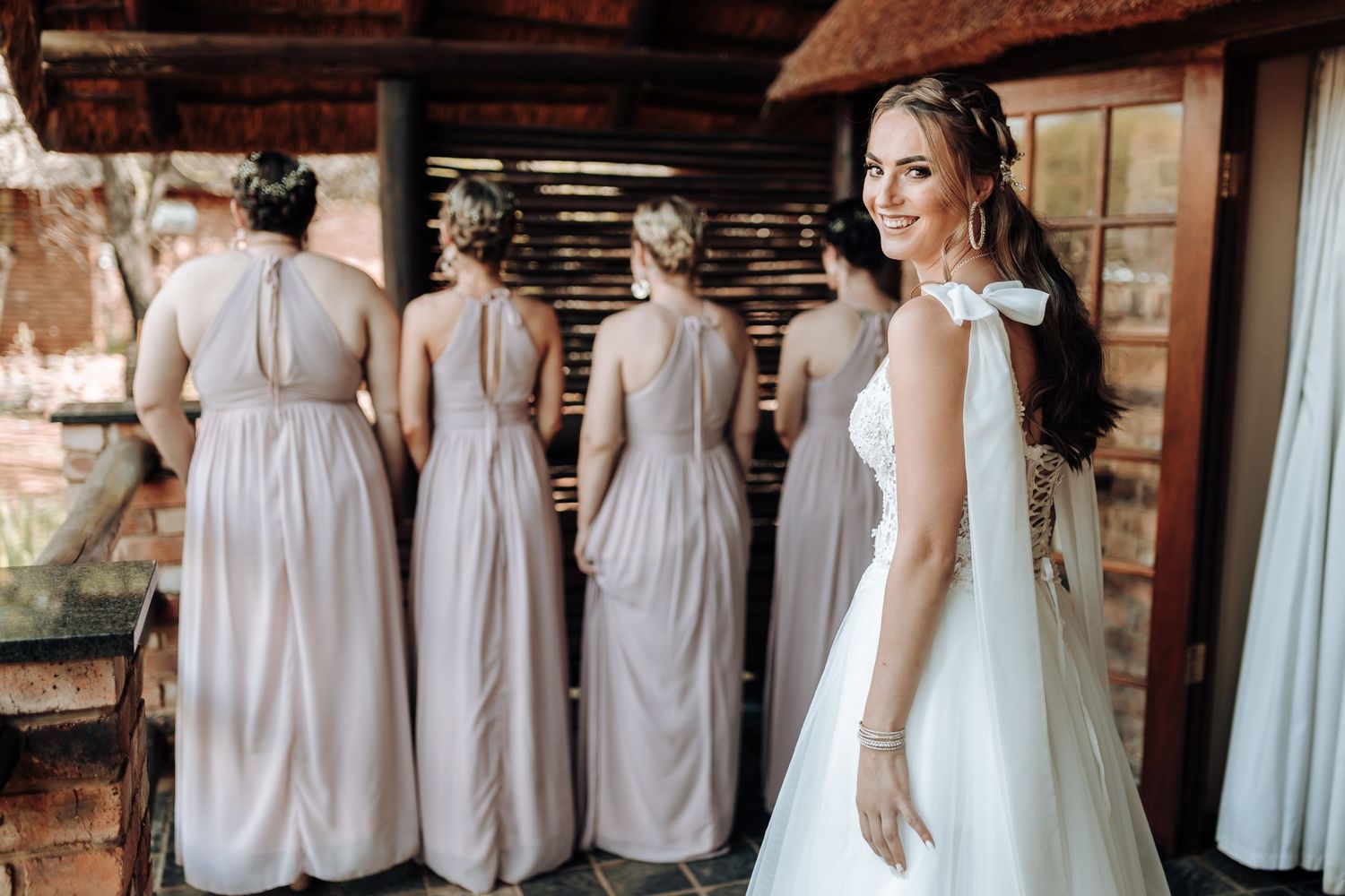 Bridesmaids in pale pink dresses stand together while a bride in white looks on in a rustic venue.