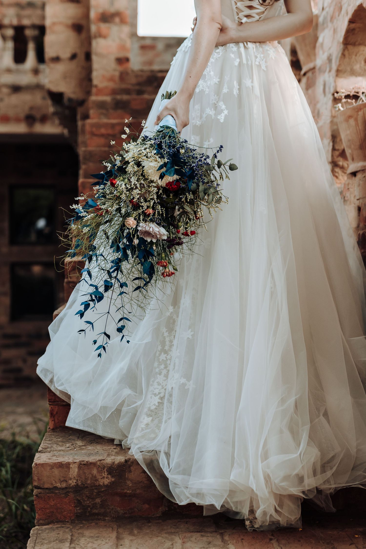Detail shot of white wedding gown and dramatic cascading blue floral bouquet.