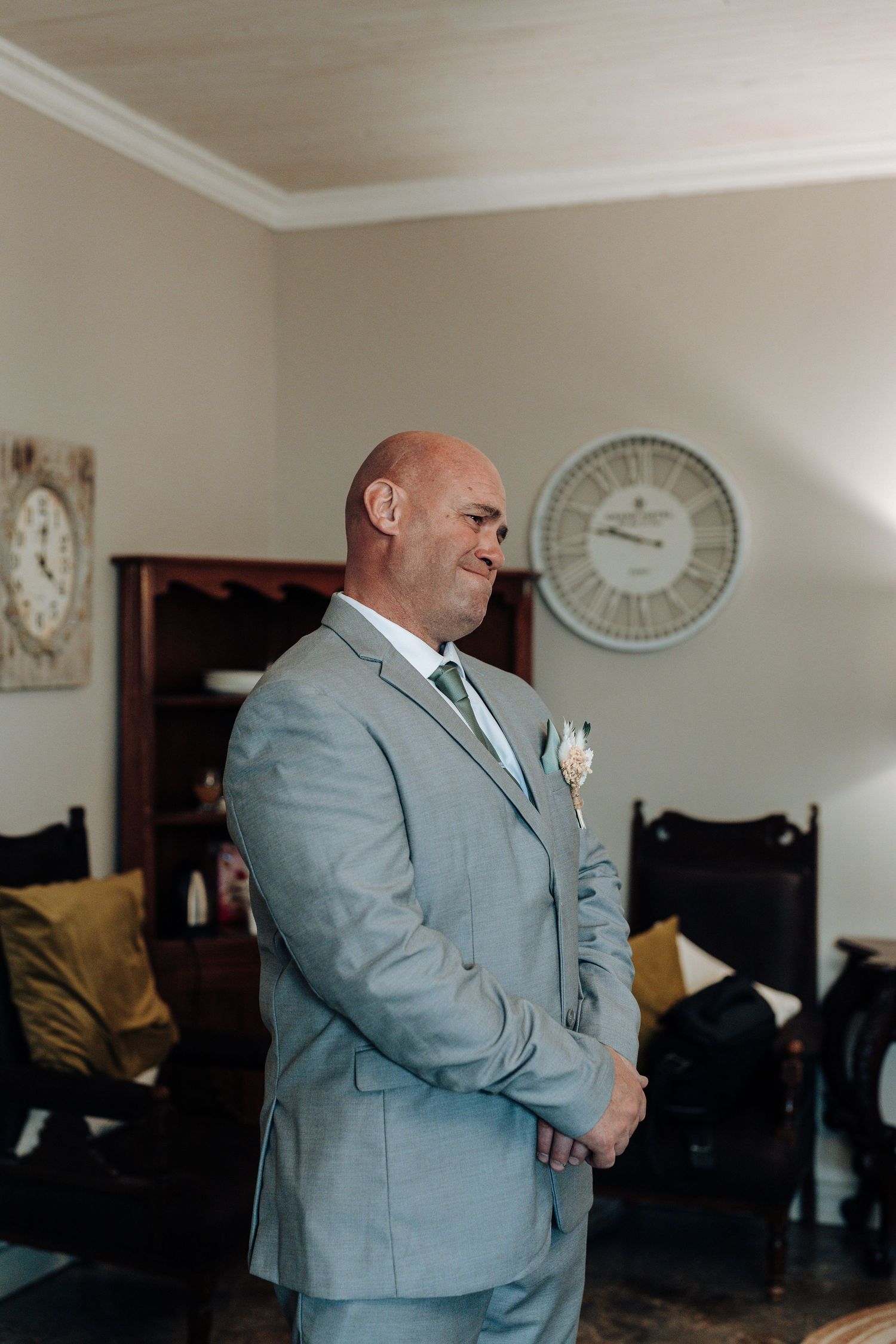 Groom in light grey suit stands emotionally in preparation room with vintage clocks on wall.
