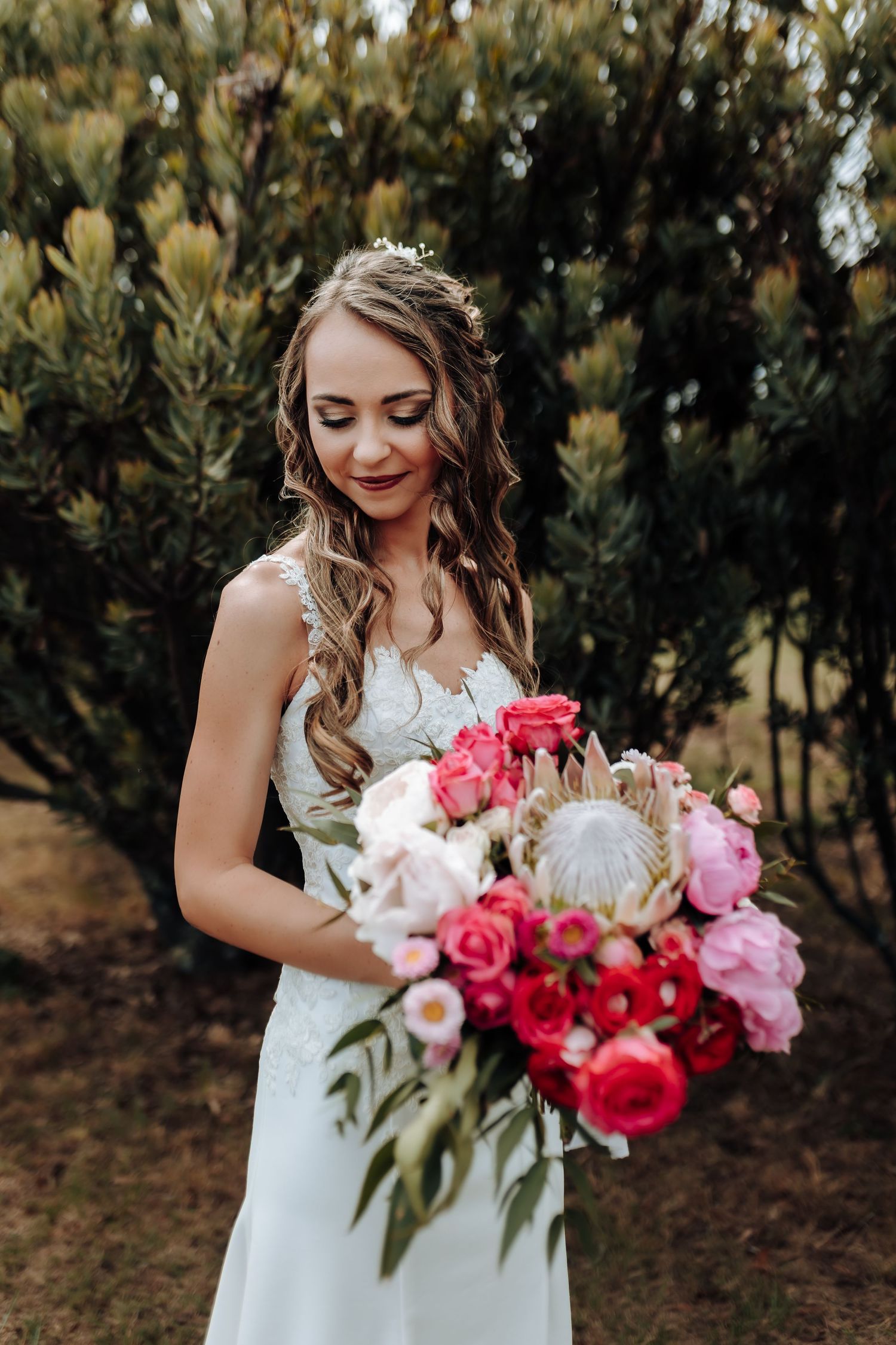 Close-up of bride holding luxurious bouquet with roses and protea flowers against greenery backdrop.