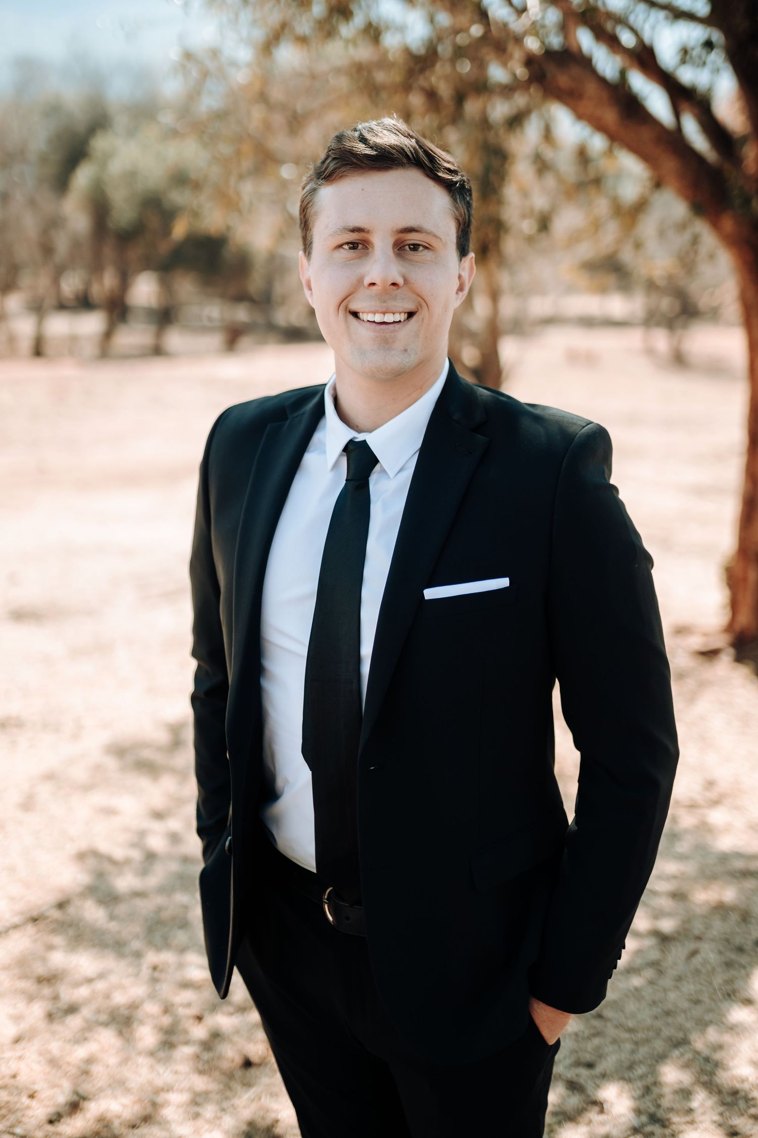 A smartly dressed person in a black suit and tie smiles warmly in an outdoor portrait setting.