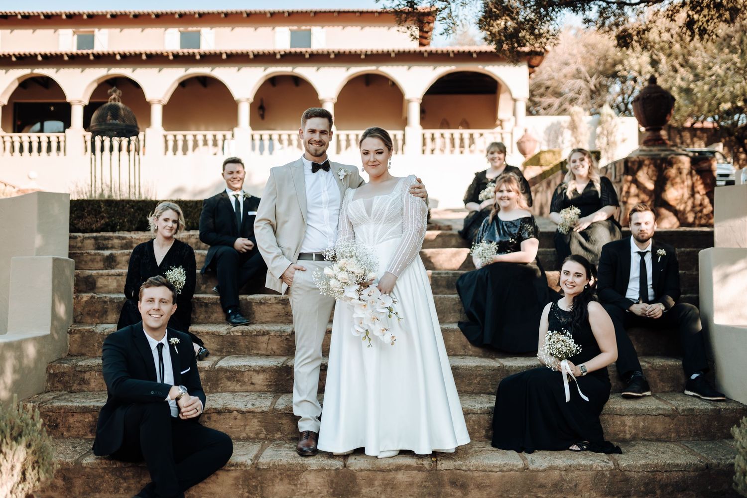 Wedding party poses on stone steps of a Spanish-style building with arched colonnade.