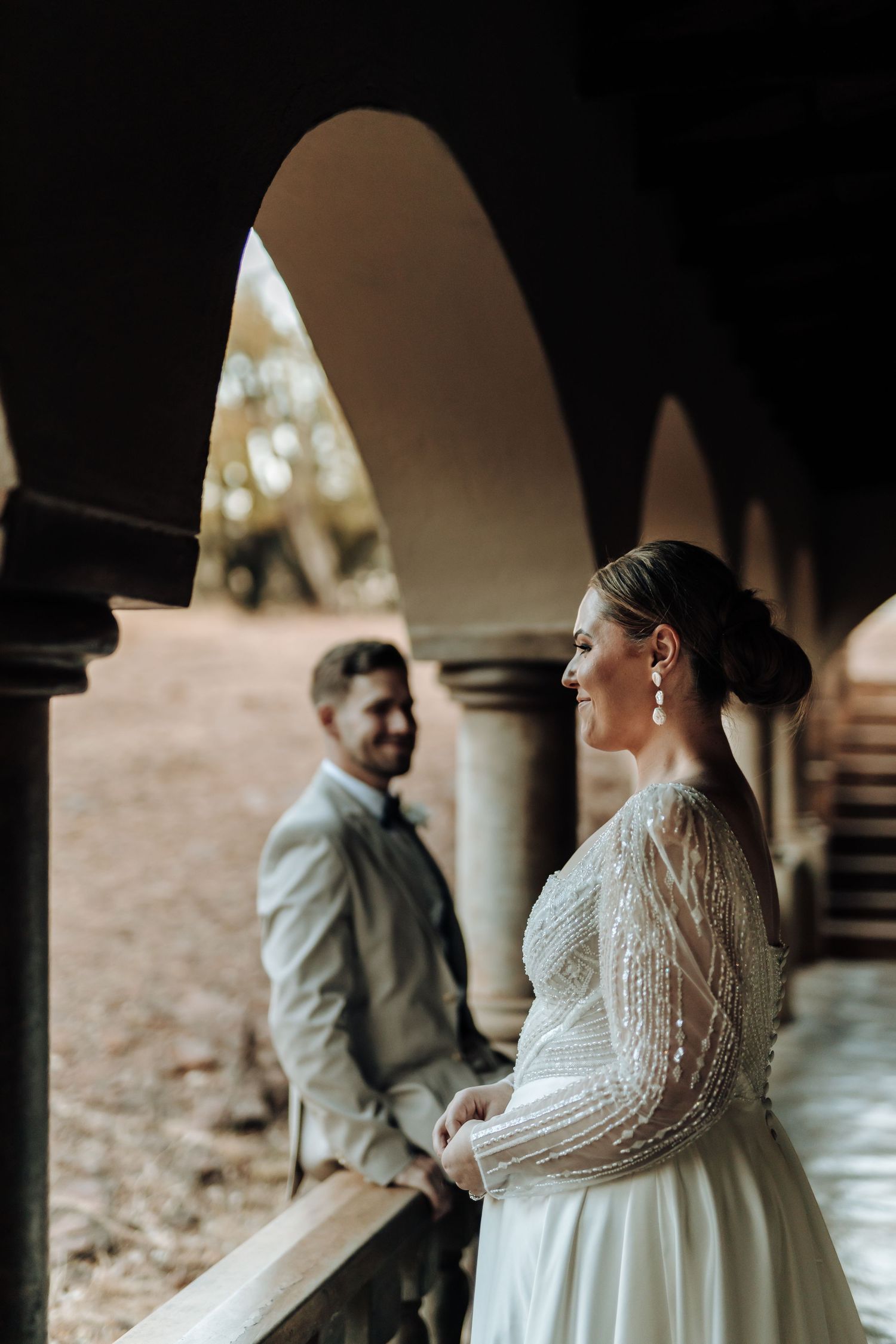 Wedding couple shares an intimate moment beneath ornate stone archways in a romantic architectural setting.