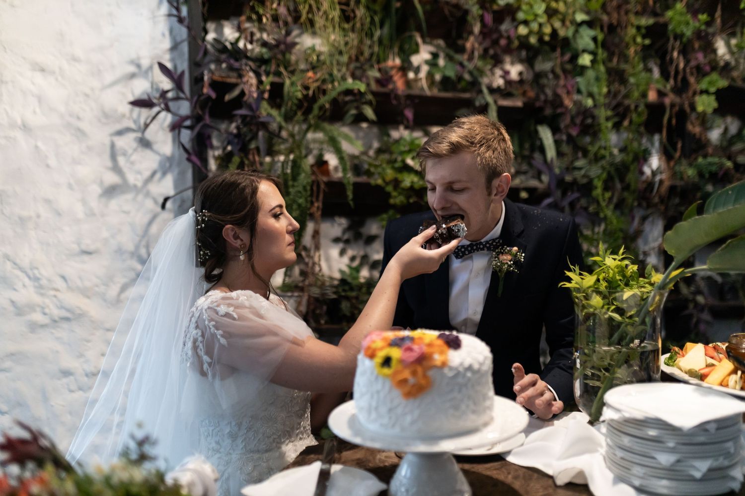 Bride and groom playfully feeding each other wedding cake at outdoor reception.