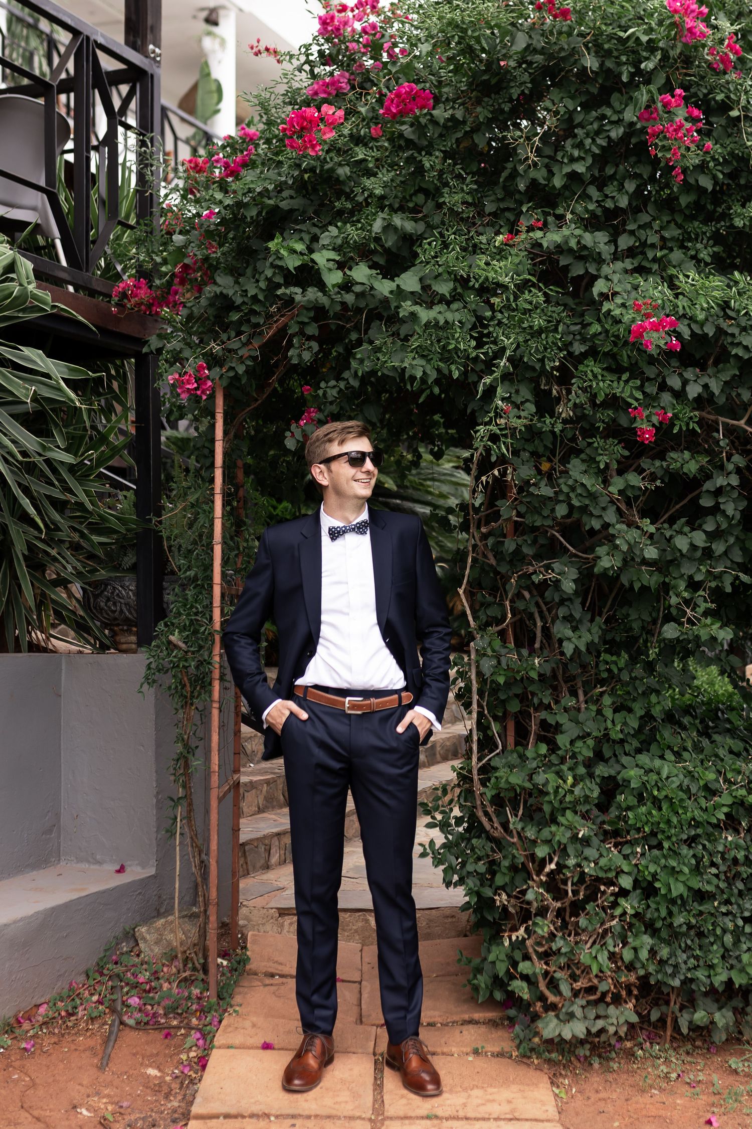 A well-dressed person poses in a navy suit against a backdrop of vibrant pink flowering bushes.