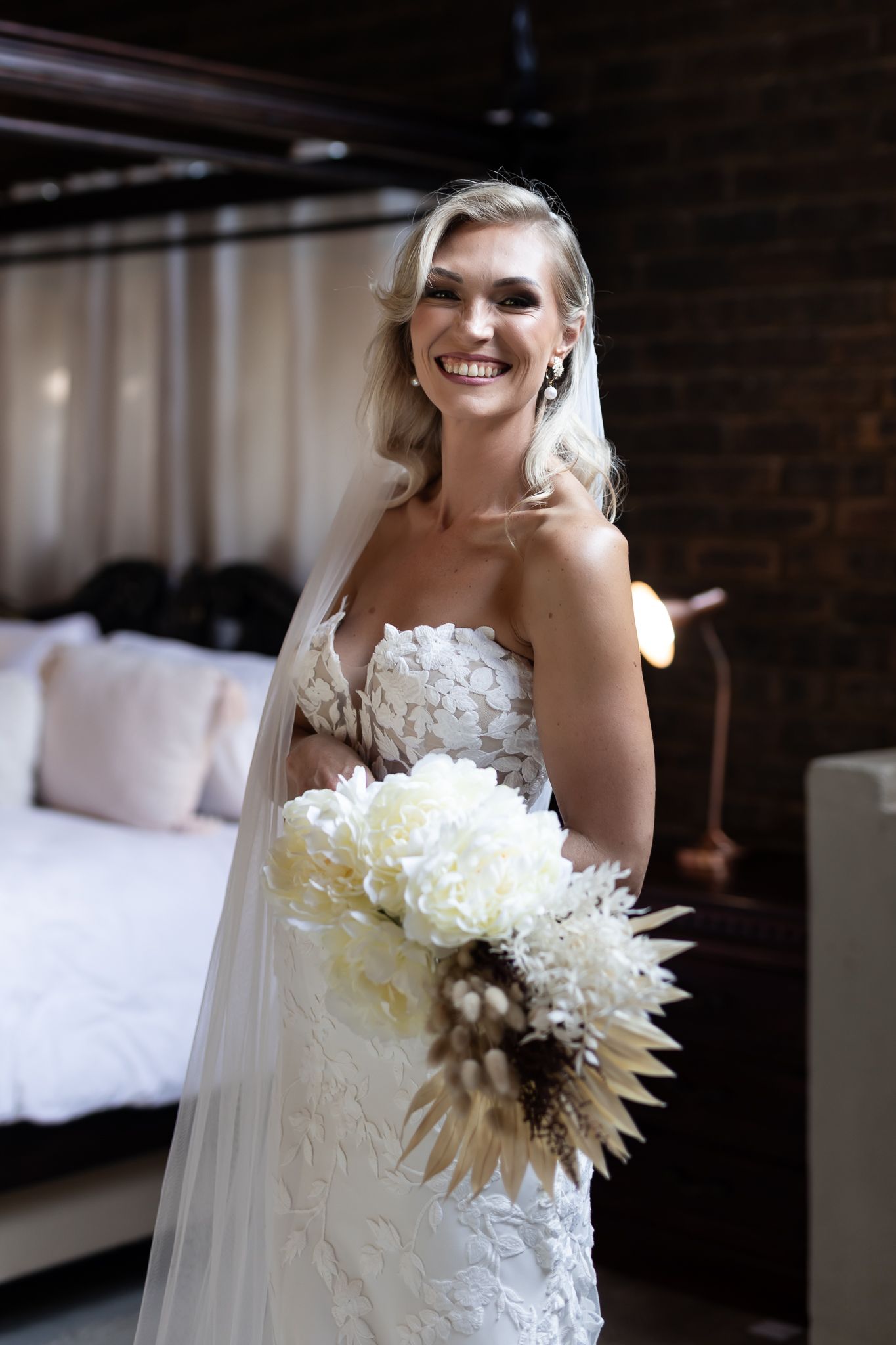 Candid moment of bride in strapless lace gown holding modern white and dried flower bouquet.
