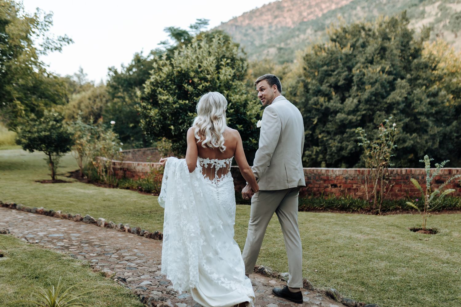 Couple walking along garden path with mountain view in elegant wedding attire.