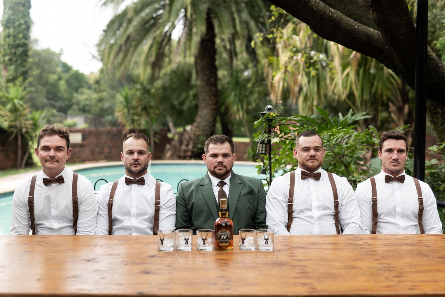 Groomsmen in suspenders and bowties sit at a wooden table with a bottle near a pool.