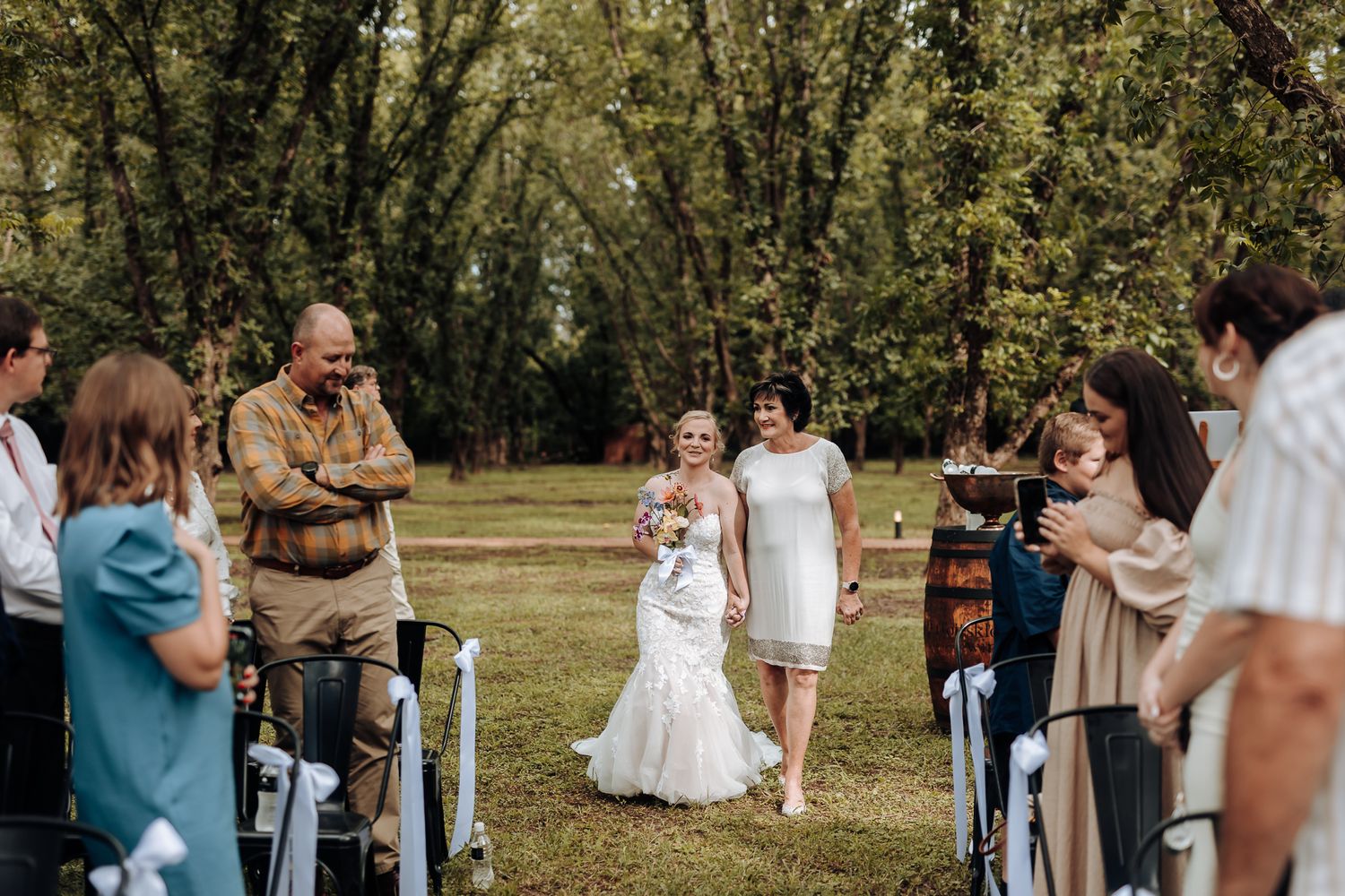 Wedding processional in a wooded outdoor venue with guests seated along the aisle.
