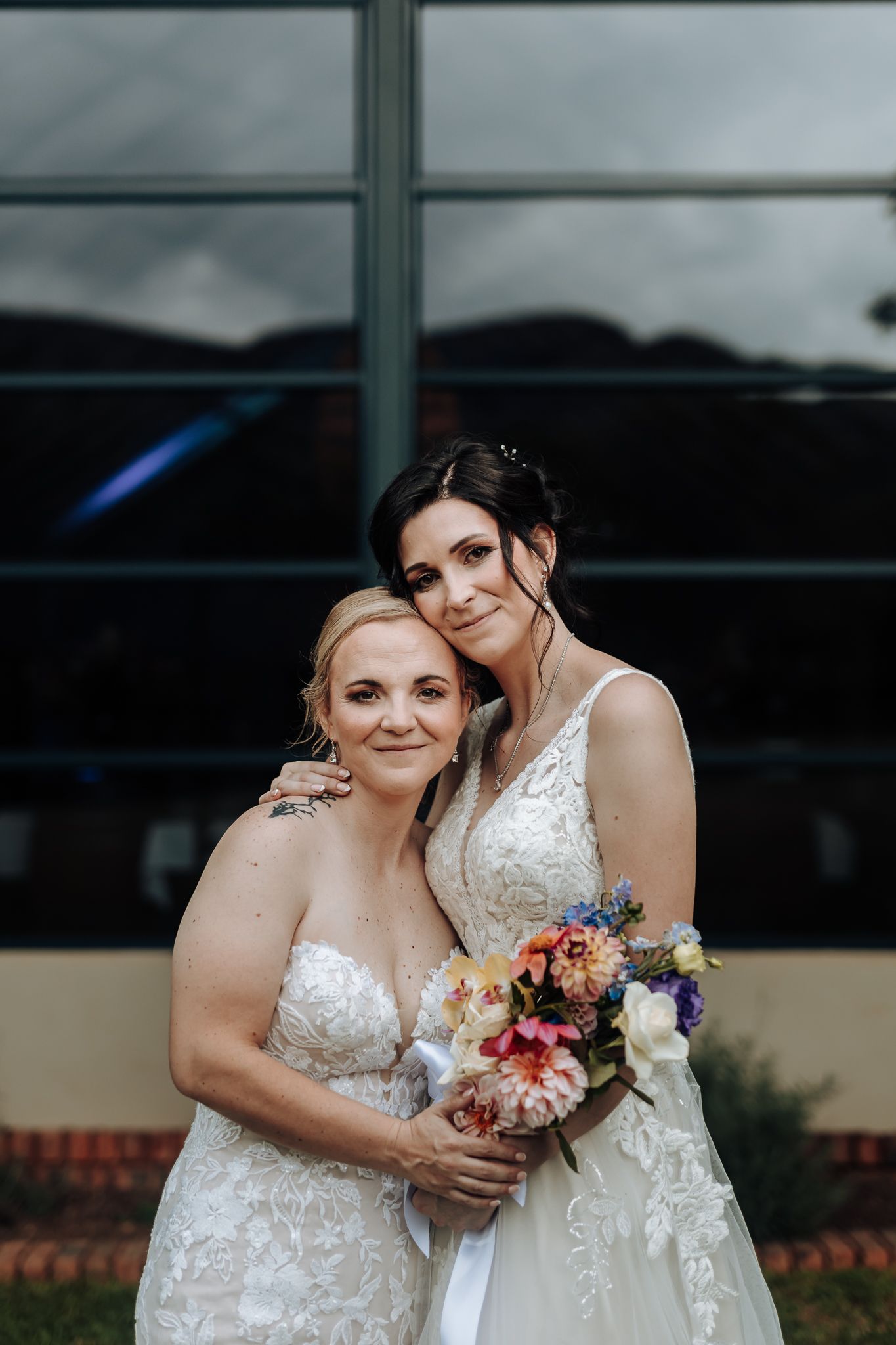 Two brides share intimate moment while holding colorful bouquets in front of large window.