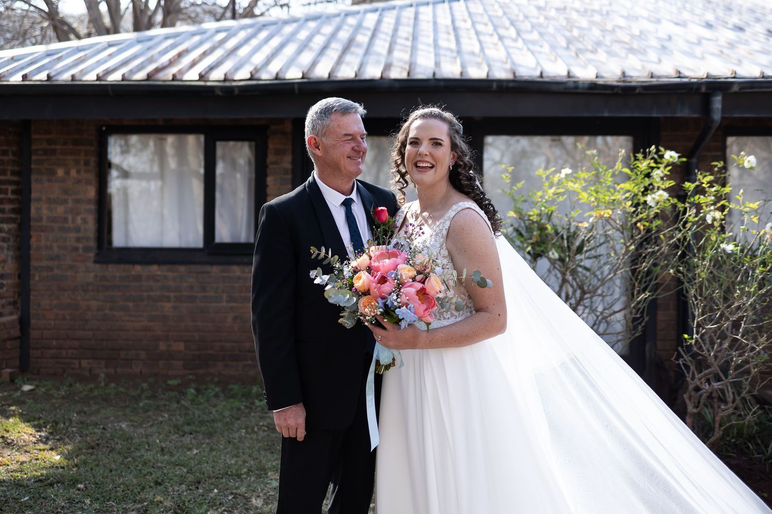 A happy couple in wedding attire pose together outdoors, with the bride holding a vibrant pink and white bouquet.