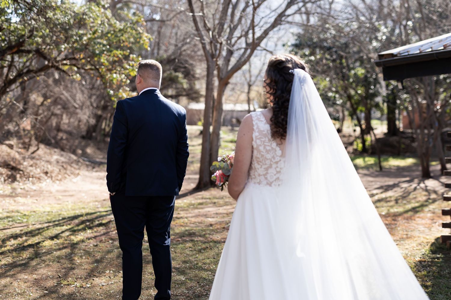 A bride in a flowing white dress and veil walks with her groom in a dark suit along an outdoor wooded path.