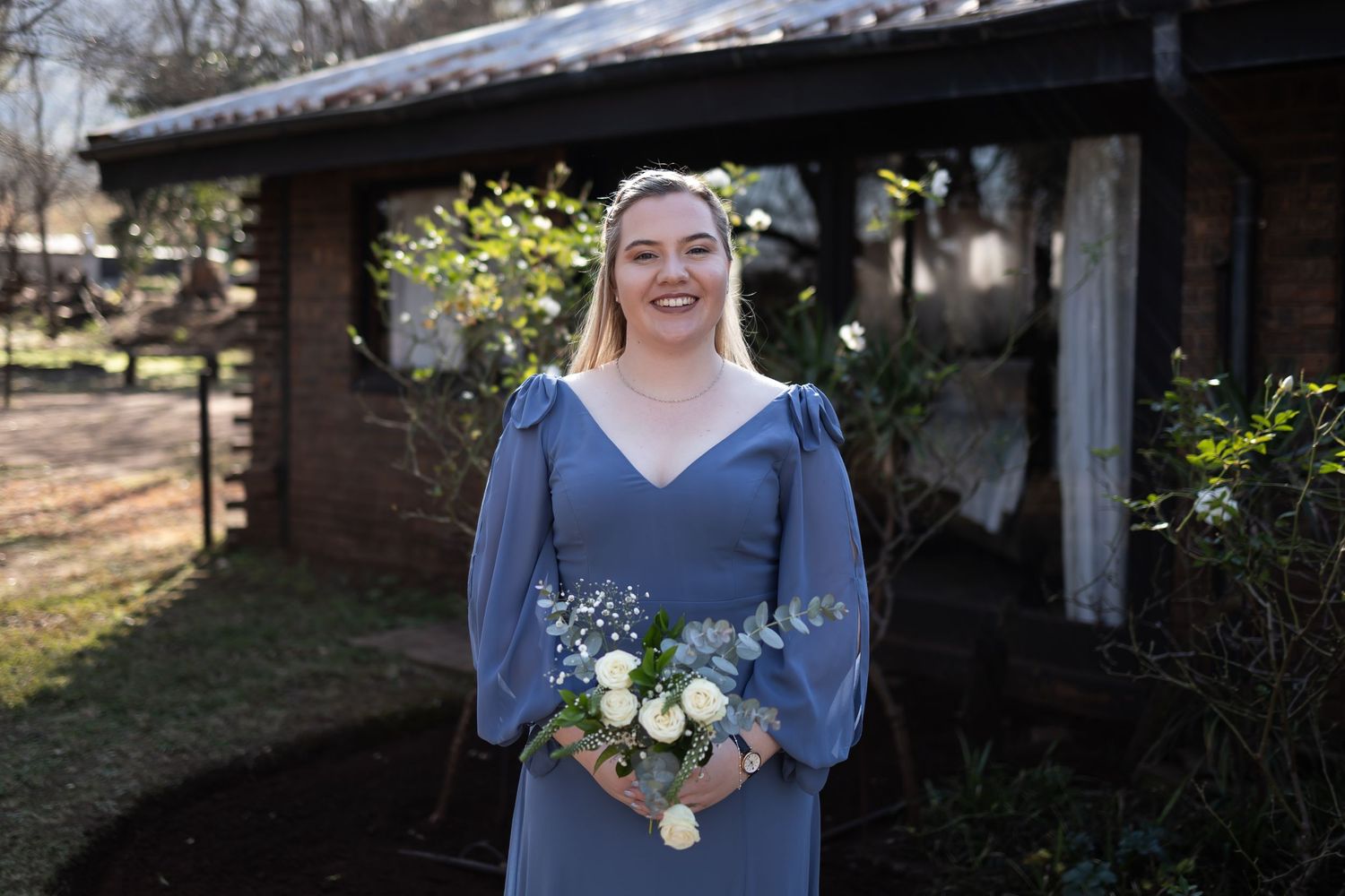 Person in dusty blue bridesmaid dress holds delicate white and green floral bouquet during outdoor wedding celebration.