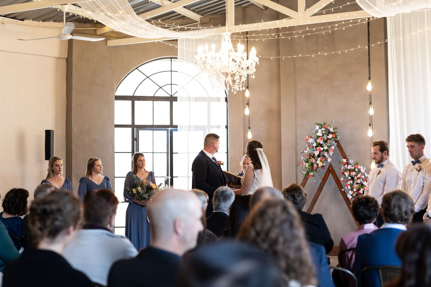 Indoor wedding ceremony at an elegant venue with arched windows, chandelier, and romantic string lights overhead.