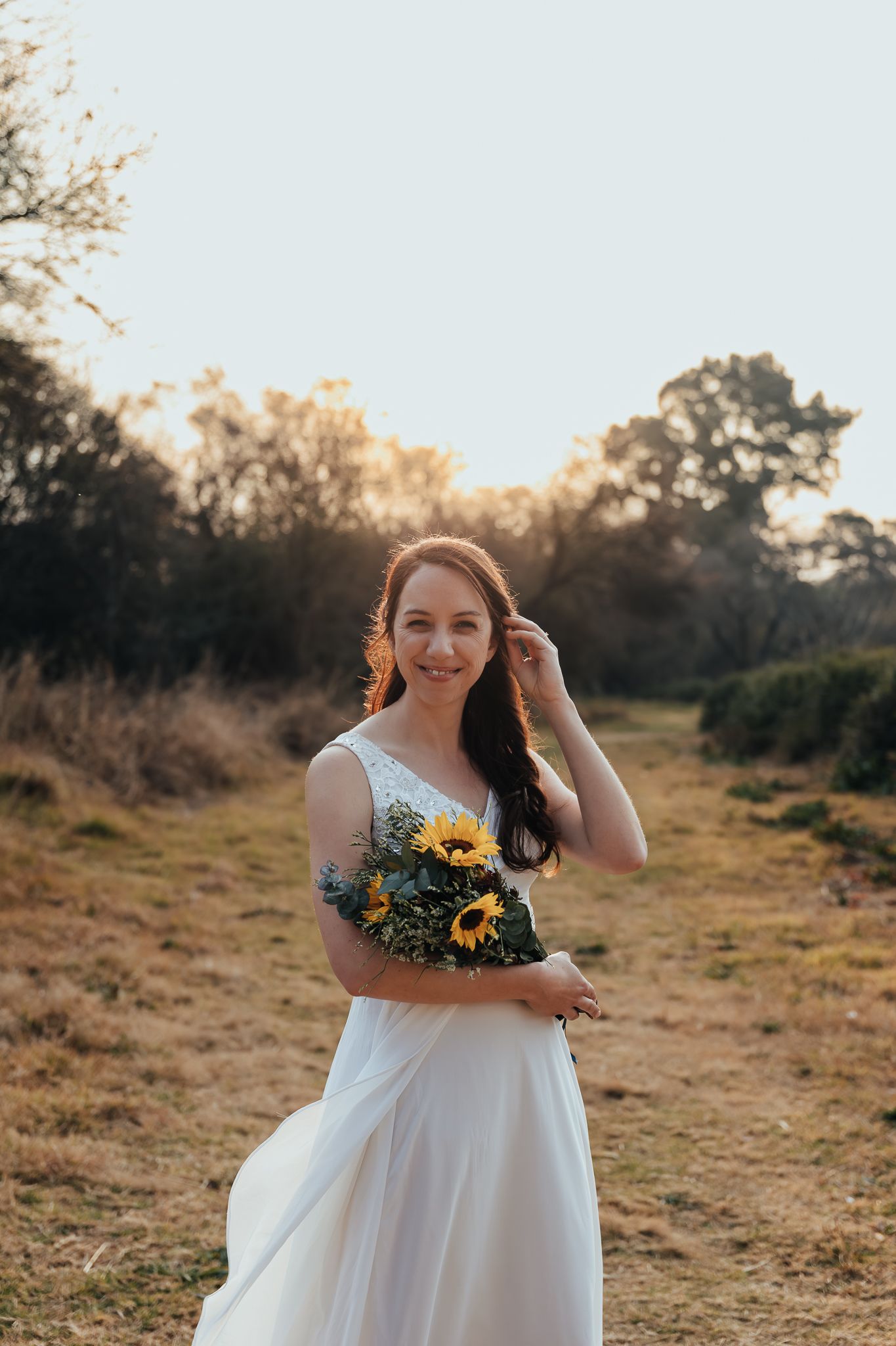 A bride in a white dress holds a sunflower bouquet while standing on a rustic trail during golden hour sunset.