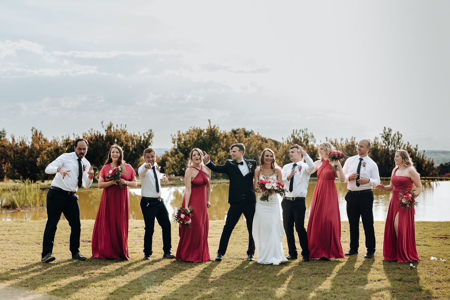 Wedding party in burgundy and white strikes fun poses against scenic lake backdrop.