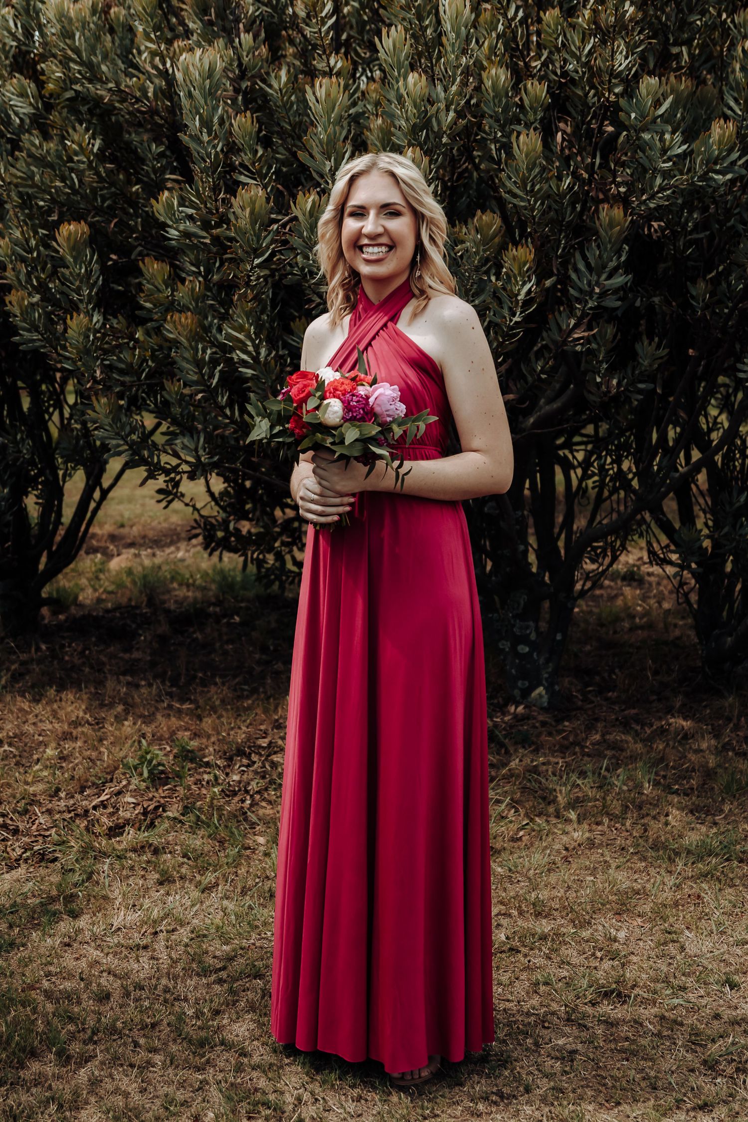 Bridesmaid in red halter gown smiles while holding floral bouquet against green foliage.
