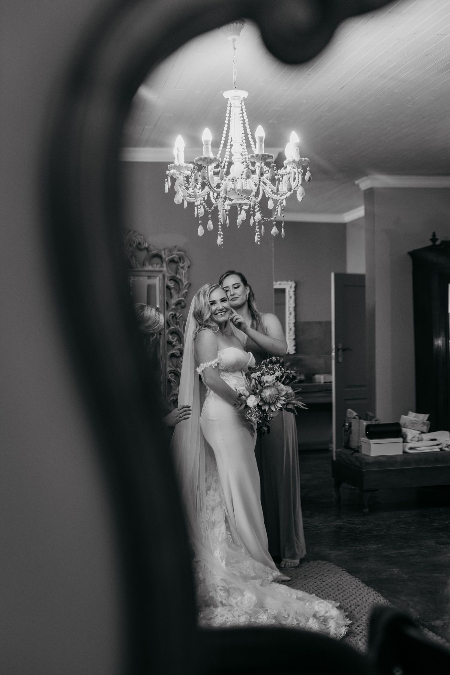 A black and white wedding photo showing a bride in a fitted gown beneath an elegant crystal chandelier.
