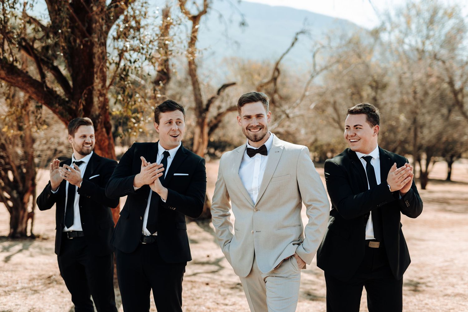 Groomsmen in black suits gesture playfully behind groom in light suit outdoors.