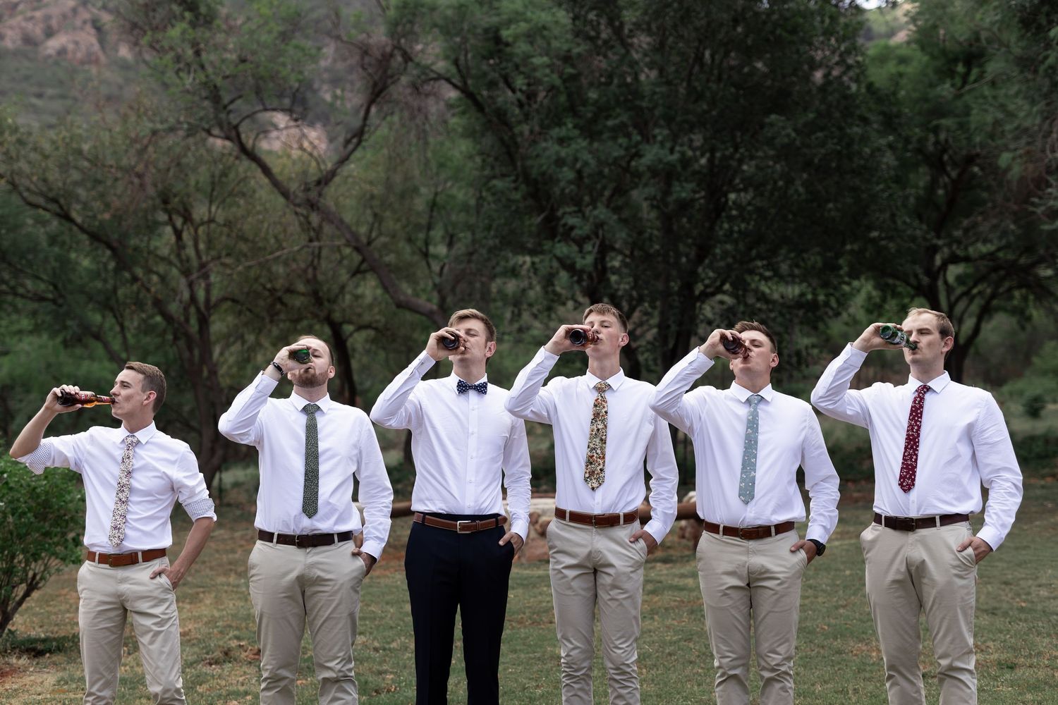 Groomsmen in light attire perform a playful pose with hands to foreheads outdoors.