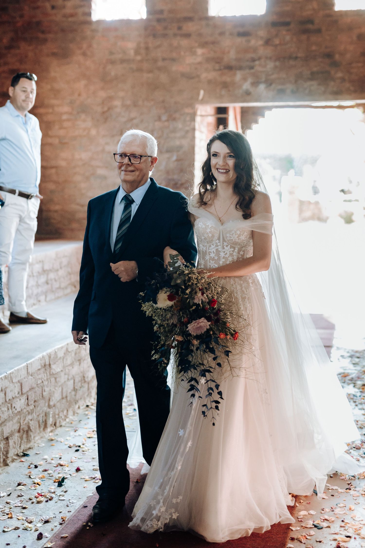 Bride in flowing white dress walking down aisle with escort at brick venue.