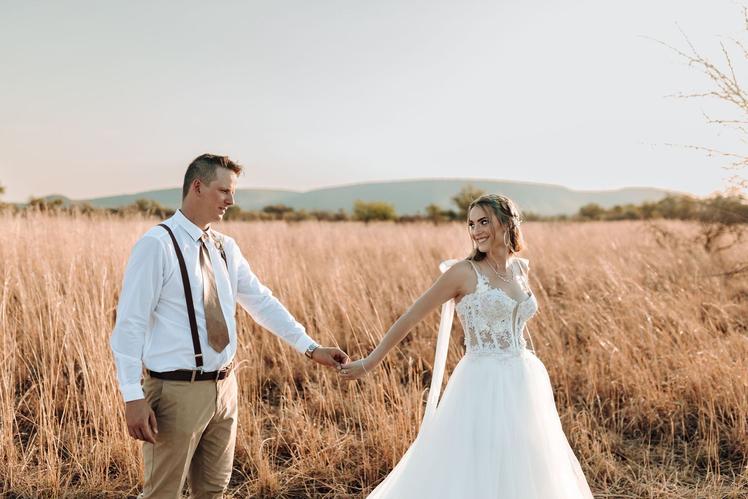 Candid moment of newlyweds walking through golden grass field at dusk.