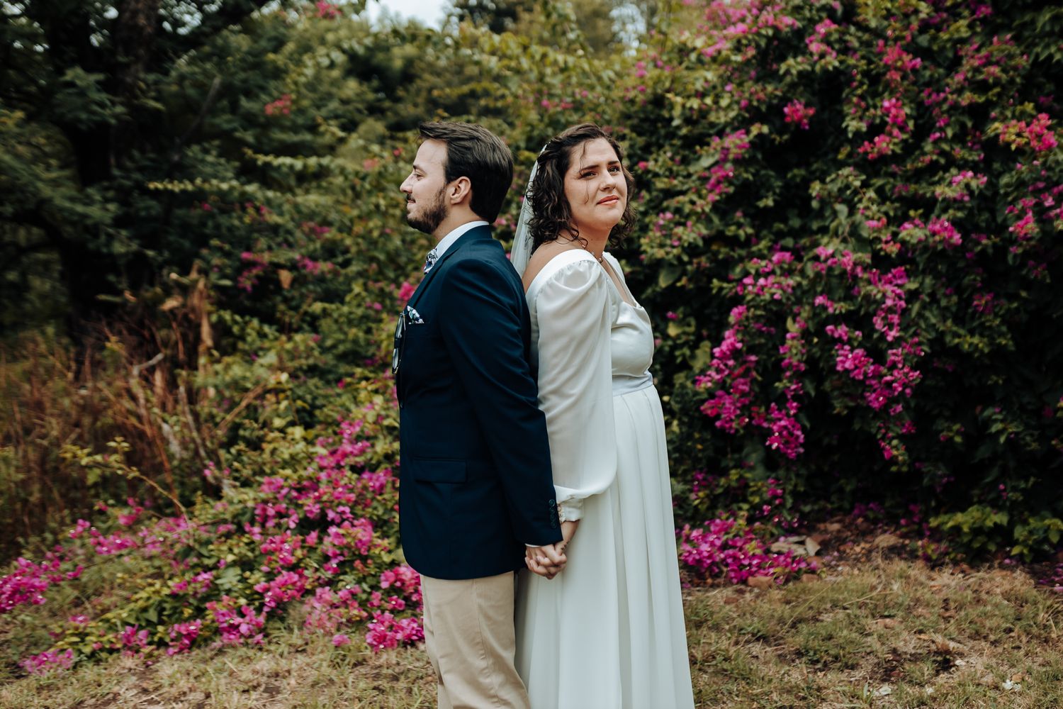 Couple in wedding attire poses back-to-back against a backdrop of pink flowering bushes.