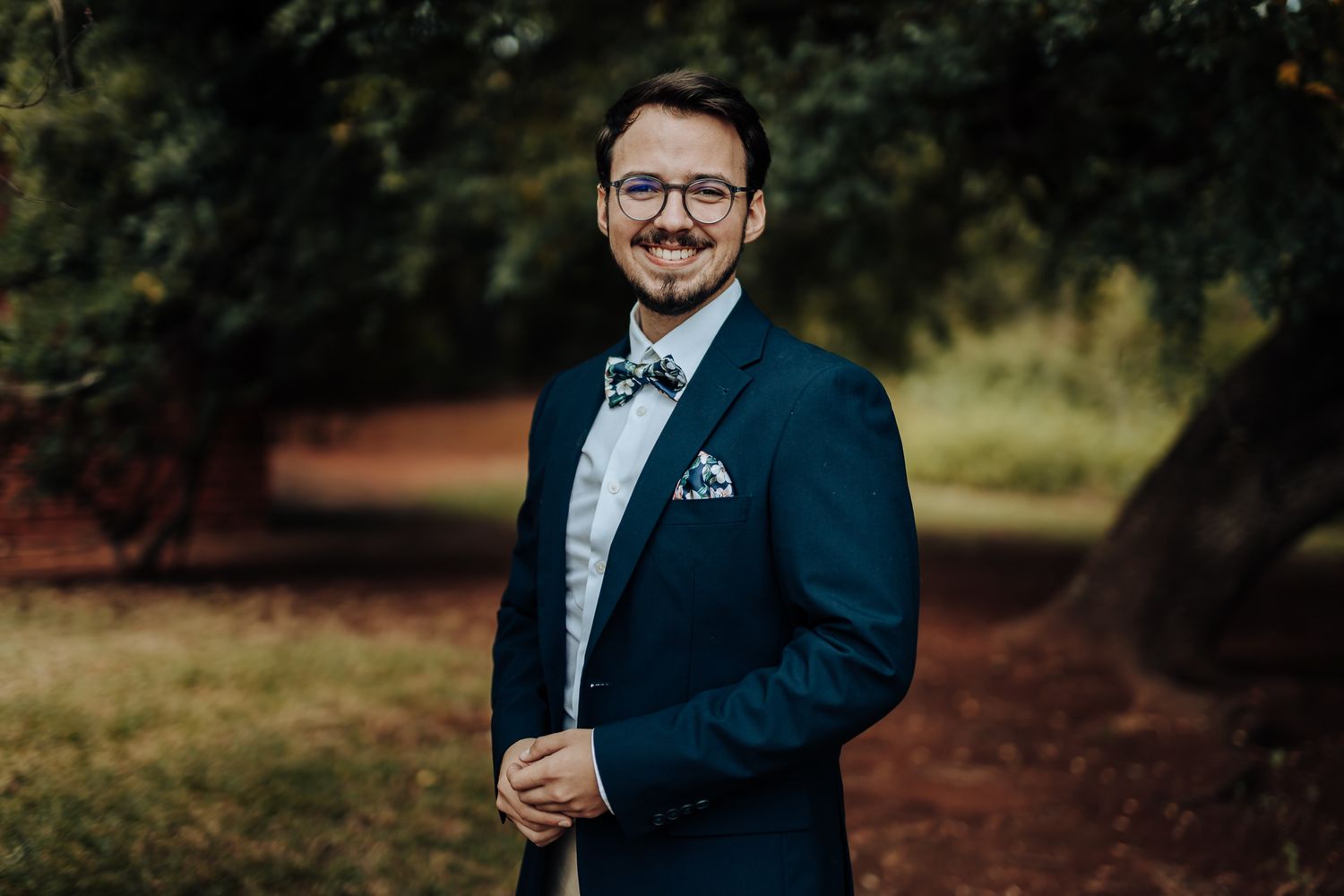 A groom wearing a navy suit and bowtie poses outdoors against a natural green backdrop.
