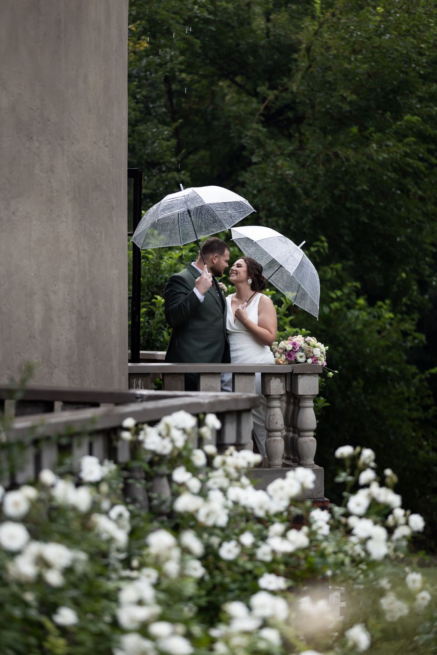 Romantic wedding couple with clear umbrellas on a stone balcony framed by white roses.