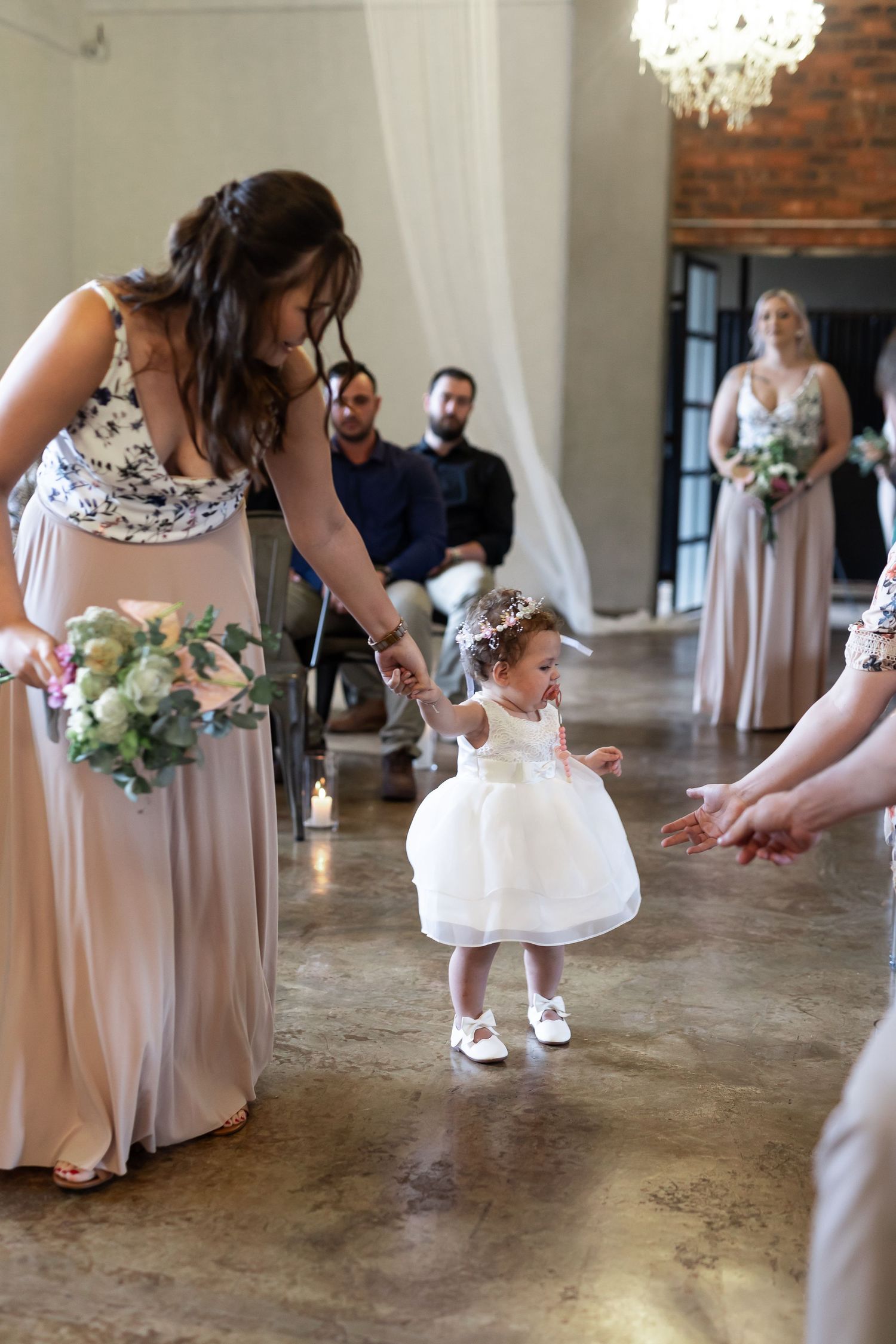 Little one in white dress taking first steps on polished concrete floor during wedding celebration.