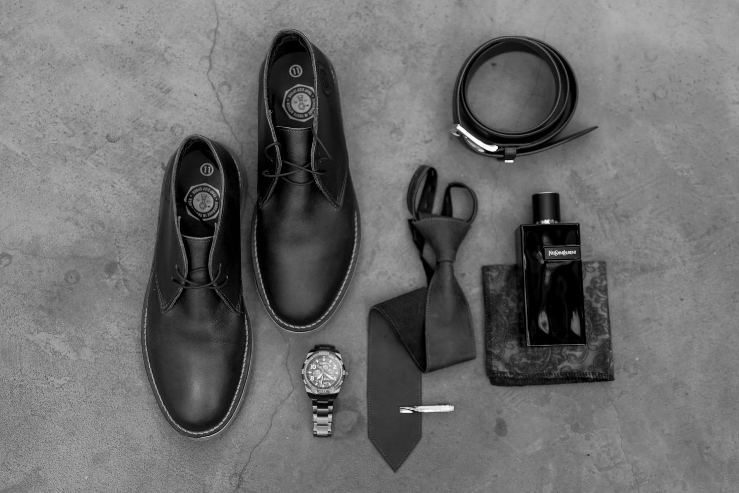 Black and white styled photo of groom accessories including shoes, watch, and cologne.