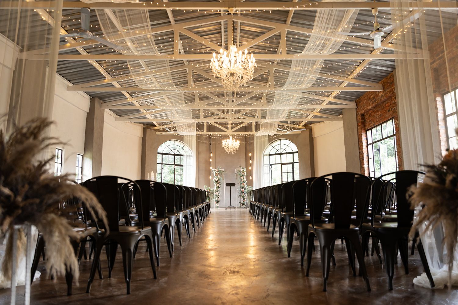 Modern wedding ceremony space with black chairs and pampas grass decor under crystal chandeliers.