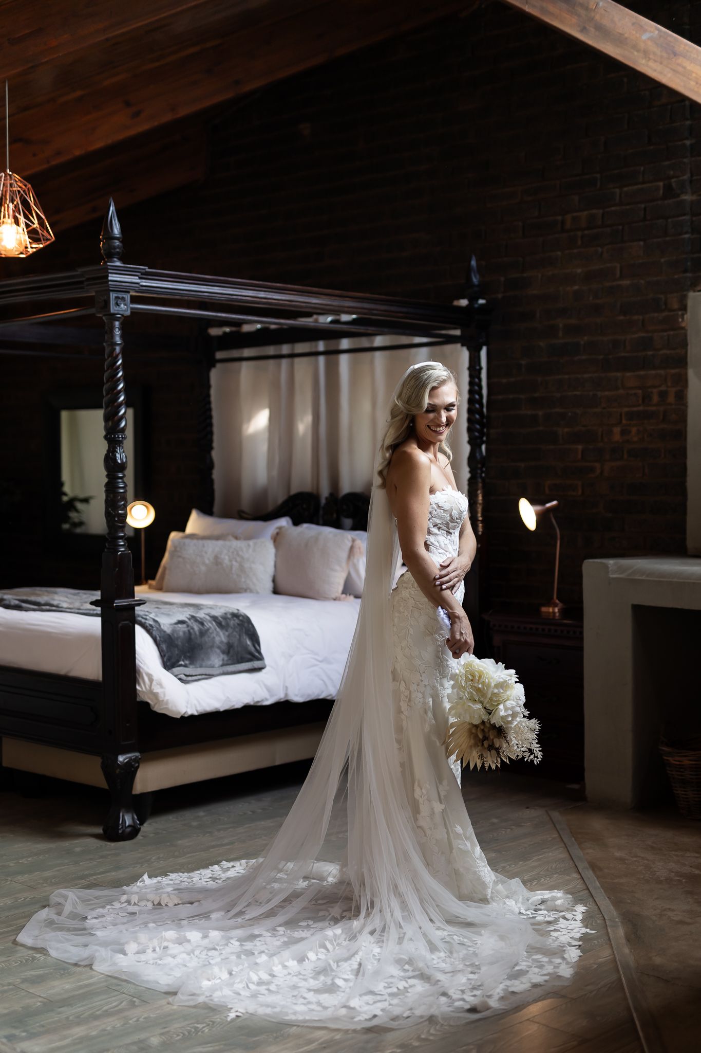 Bride in fitted lace gown holding white bouquet poses in front of vintage four-poster bed.