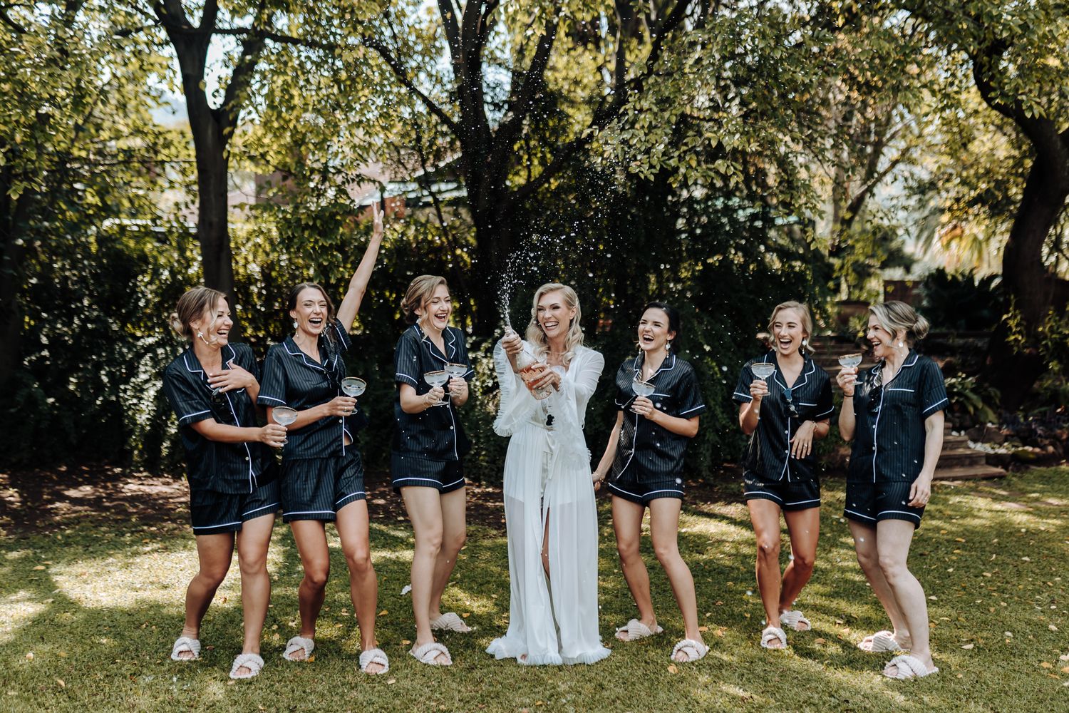 Bride and bridesmaids in matching black robes celebrating outdoors under trees.