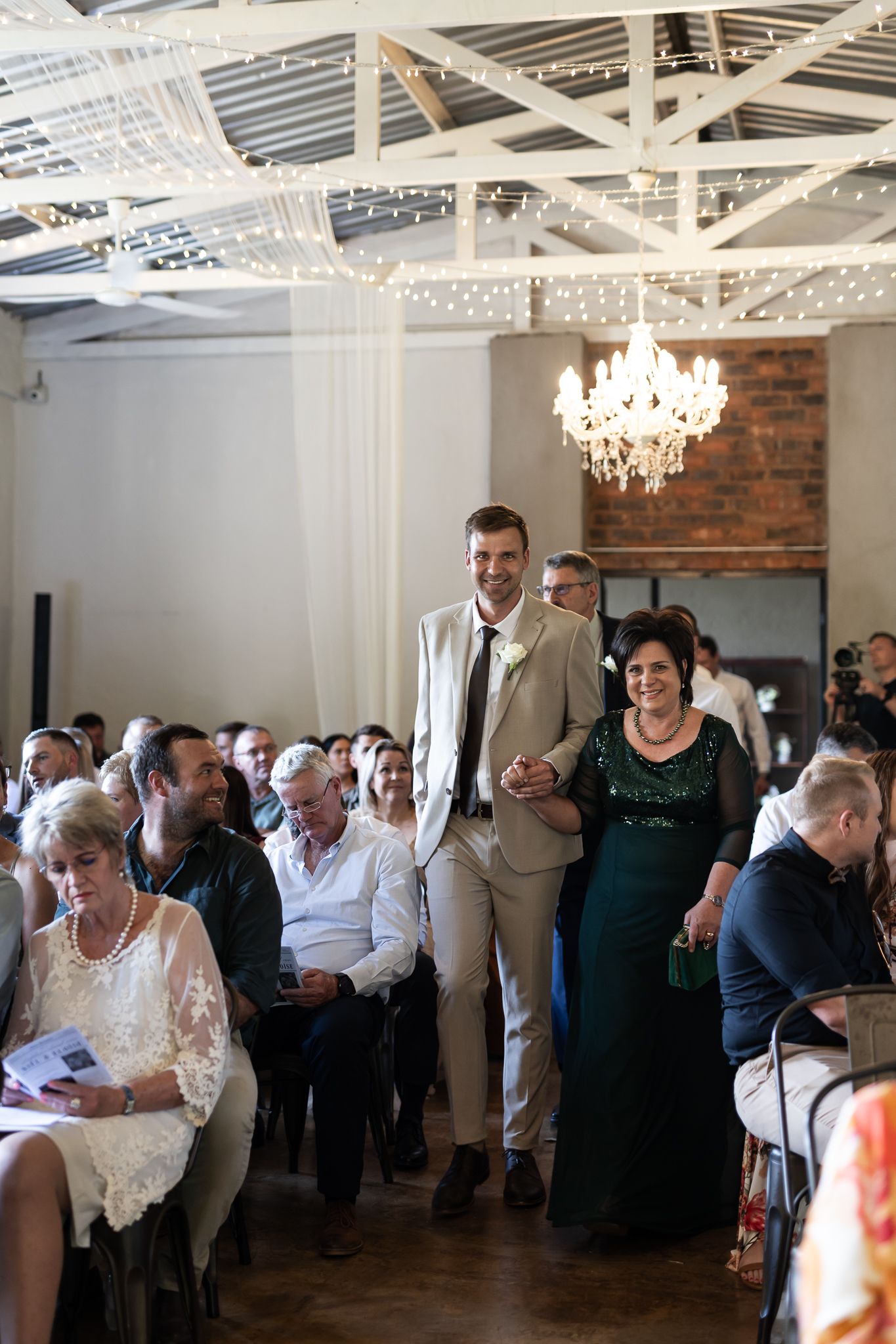Wedding ceremony guests seated in elegant venue with chandelier and exposed brick wall.