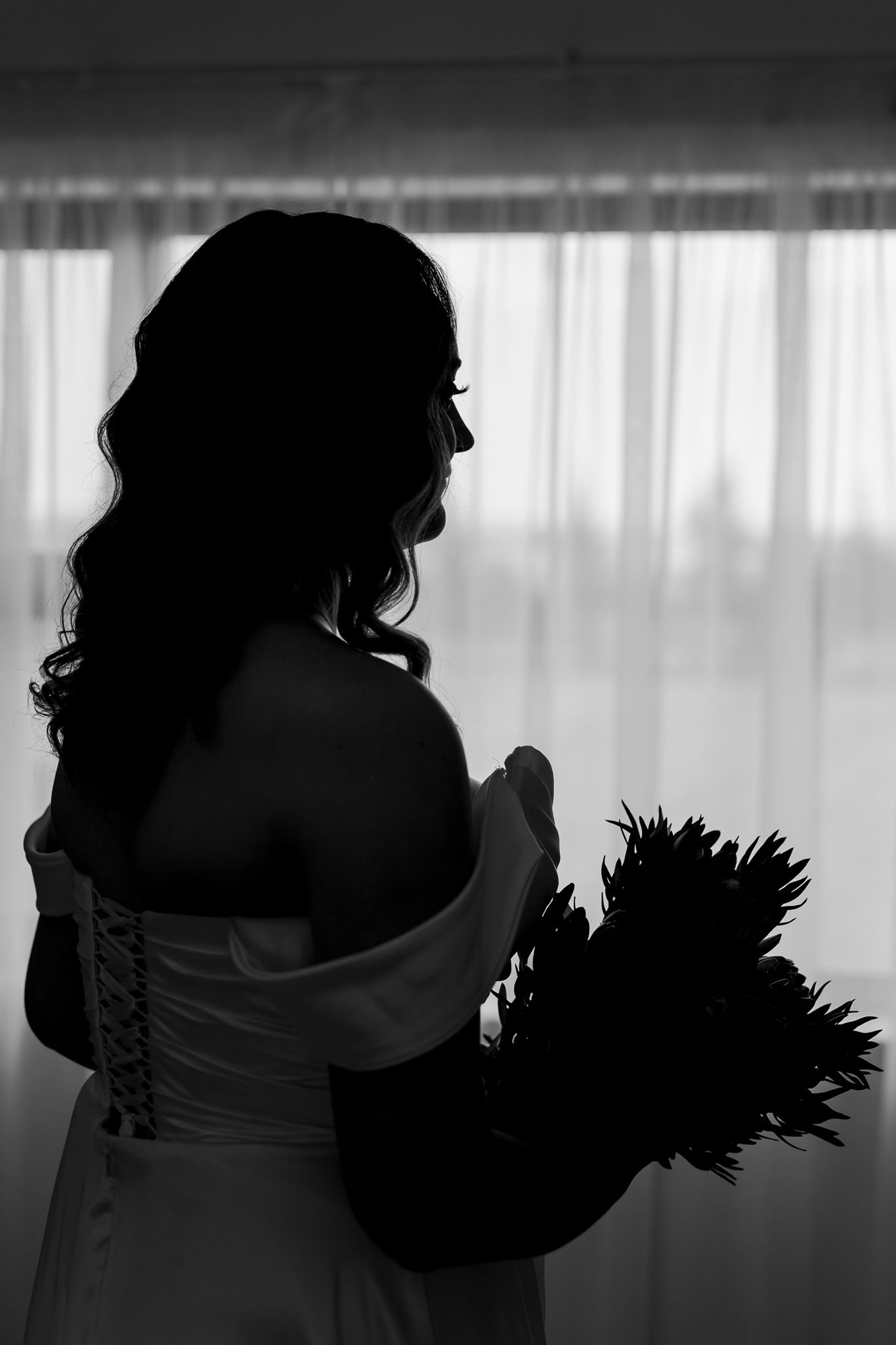 Silhouetted bride in white dress holding dark bouquet against bright window with sheer curtains.