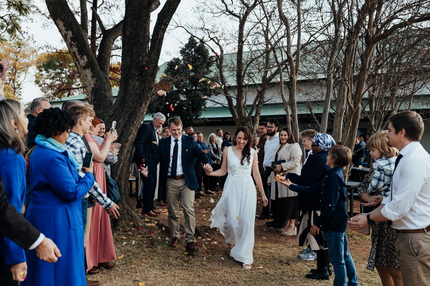 A wedding celebration taking place outdoors under large trees with guests gathered to celebrate.