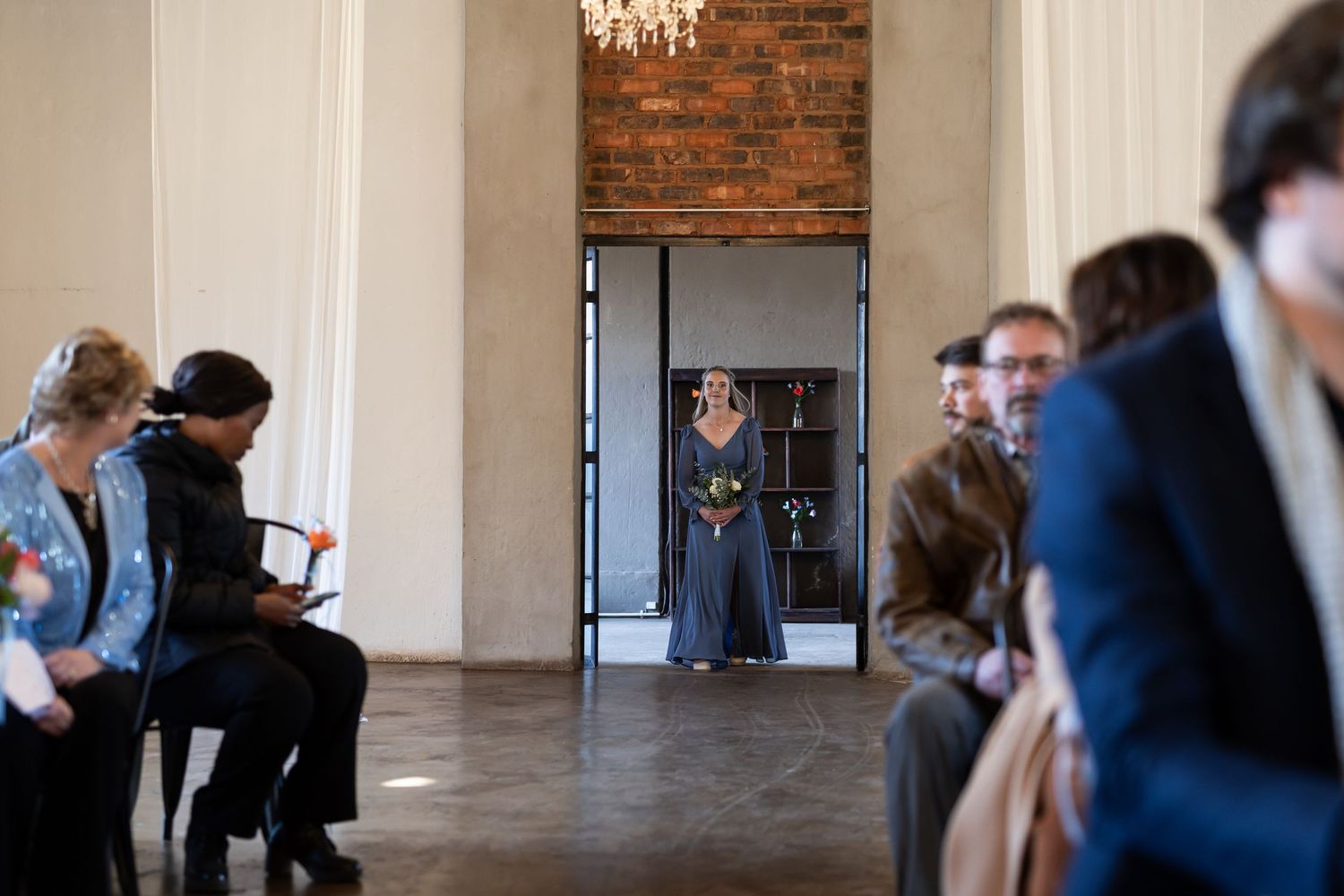 People wait in a room with exposed brick walls and white columns while someone stands in a doorway at the far end.
