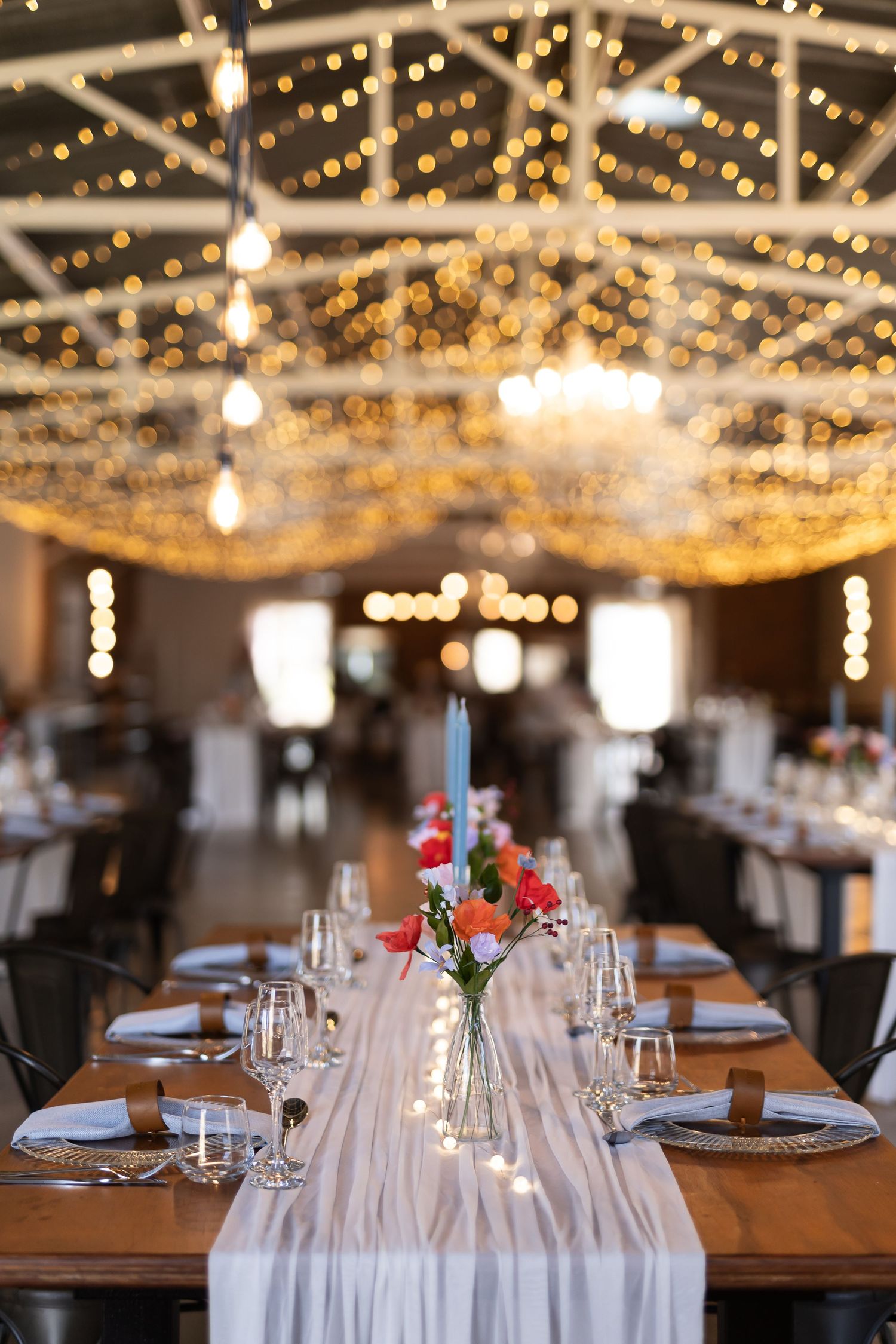 Elegant wedding reception table with white runner and floral centerpiece under twinkling fairy lights in rustic venue.
