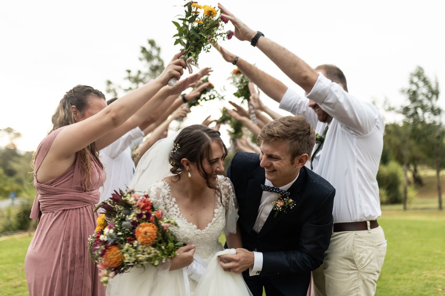 Wedding party creates celebratory arch with arms while holding flowers outdoors.