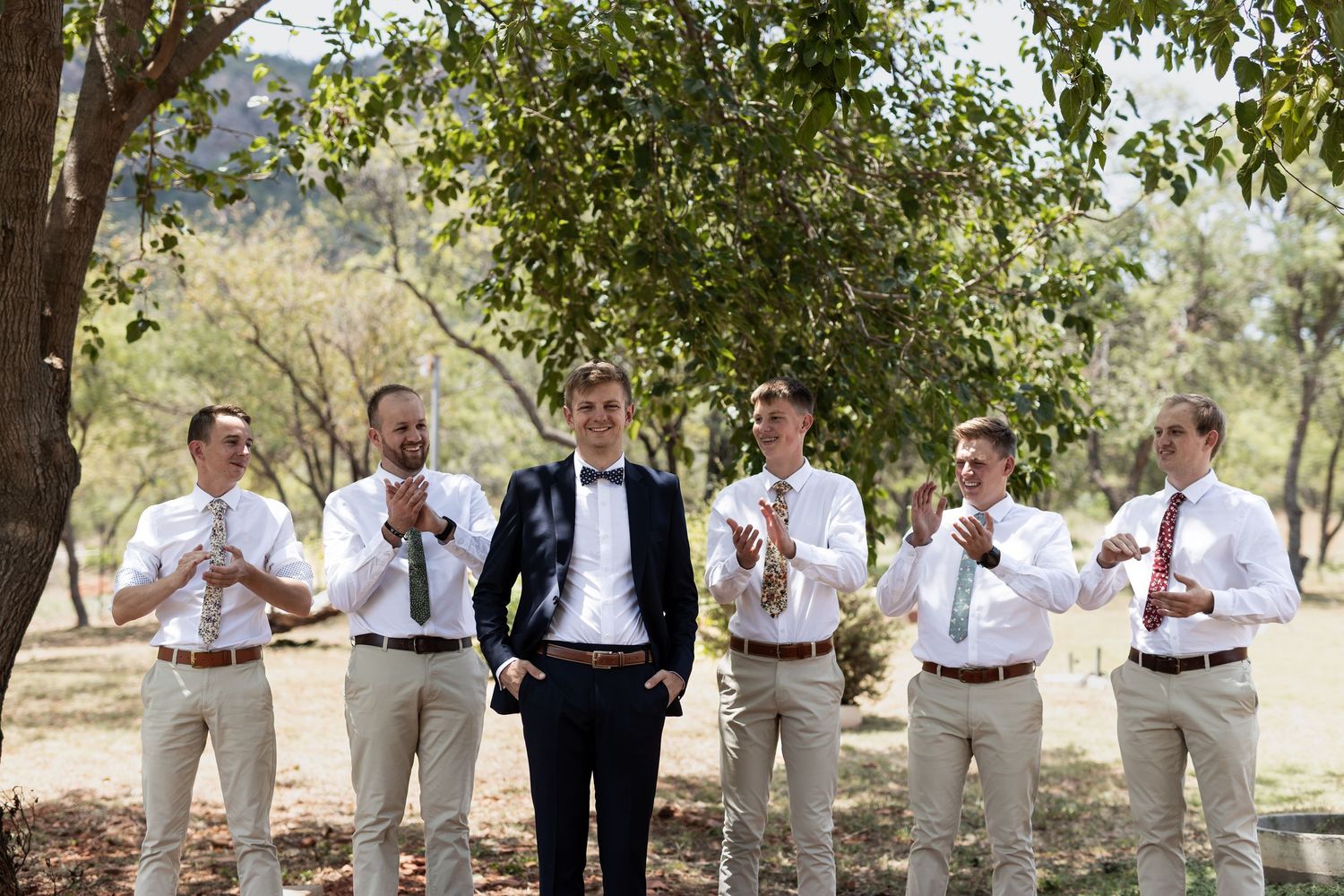 Groomsmen in khaki pants and white shirts stand alongside the groom in a navy suit under a tree outdoors.