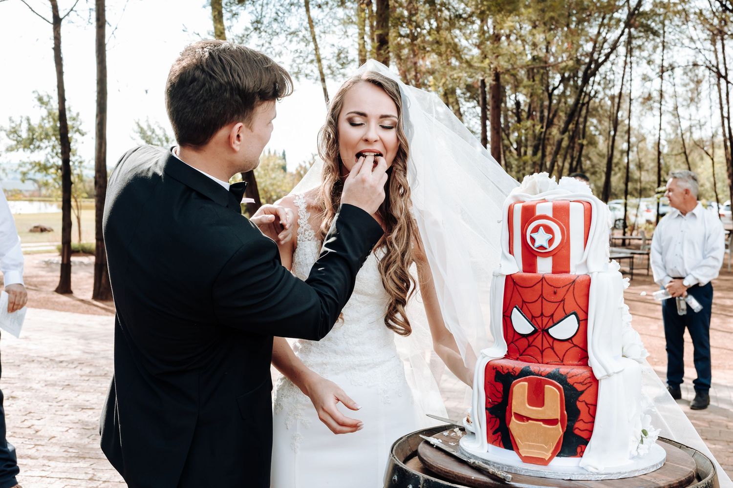 Newlyweds share cake next to a unique Iron Man themed wedding cake at an outdoor reception.